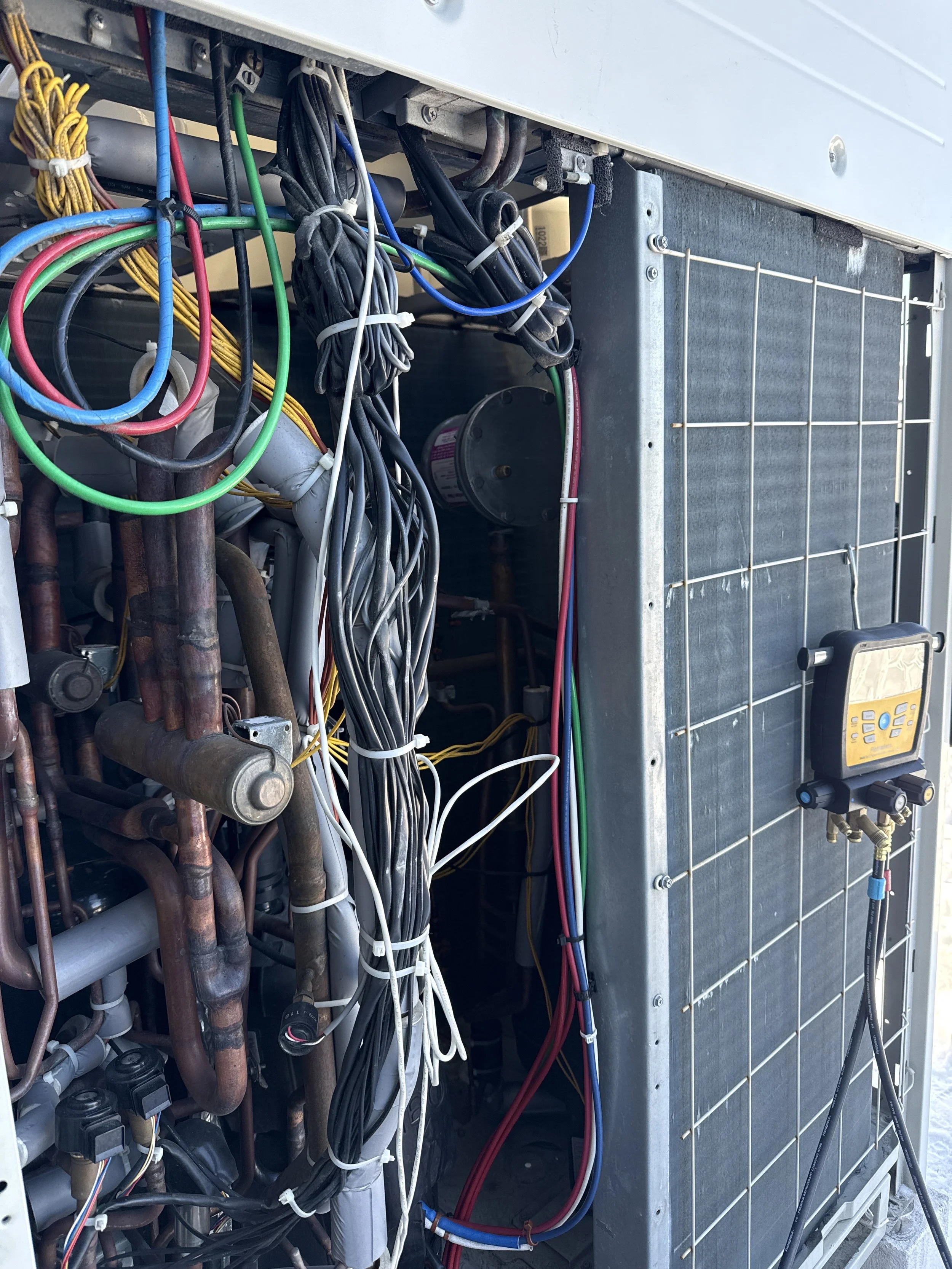 Close-up of an HVAC unit's internal wiring, including multicolored wires, electrical components, copper pipes, and a digital multimeter.