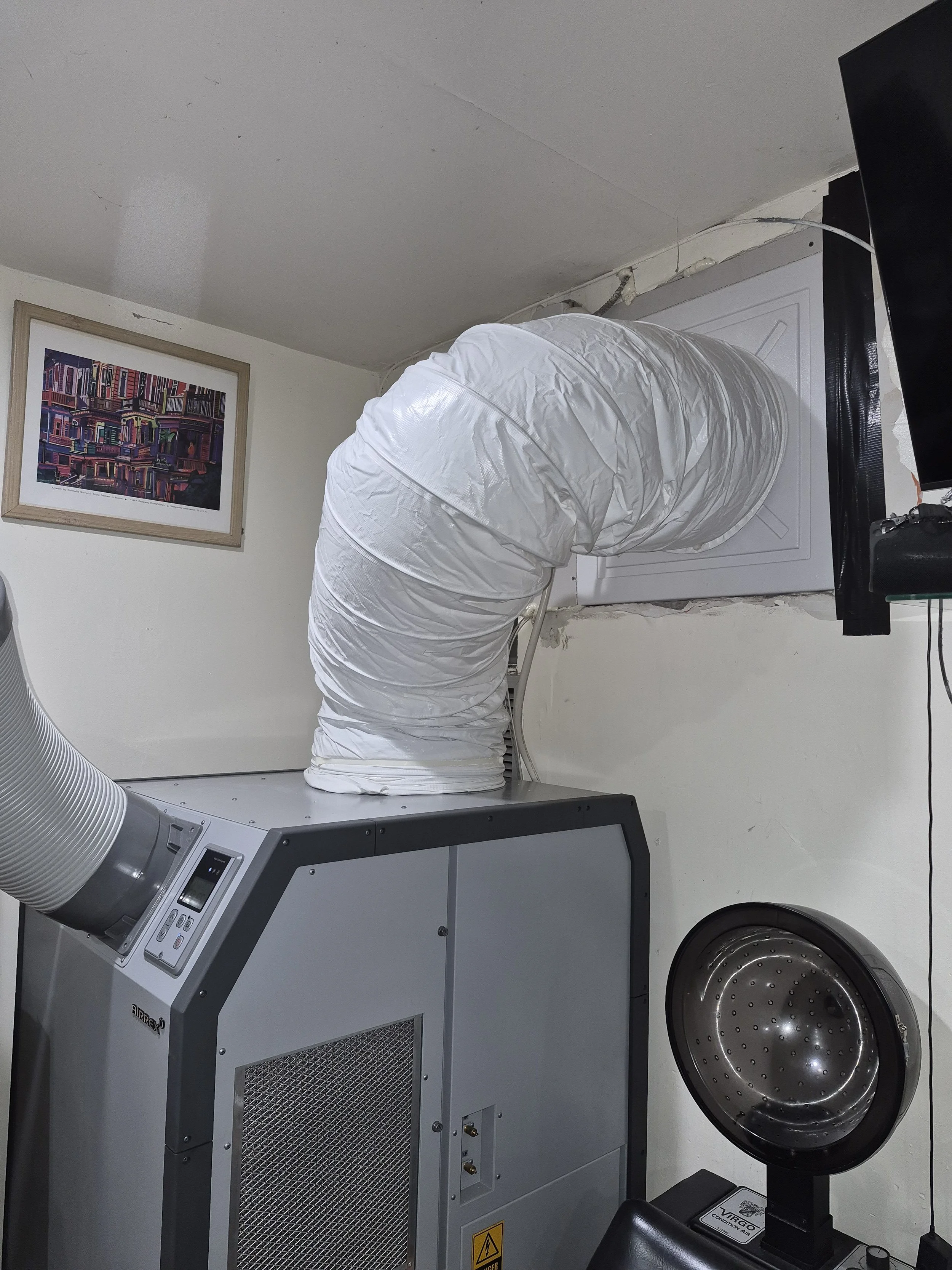 Industrial dryer with a flexible duct connected to an air vent on the wall, a framed colorful cityscape art on the wall, and a monitor on the right side.