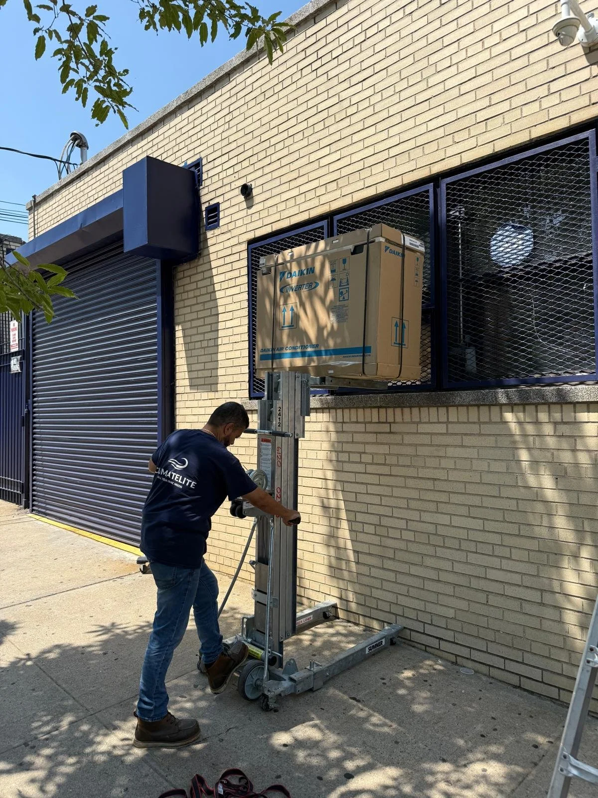 A man operating a manual lift outside a brick building, next to a box labeled for an air conditioner, with a security window and a large black garage door in the background.