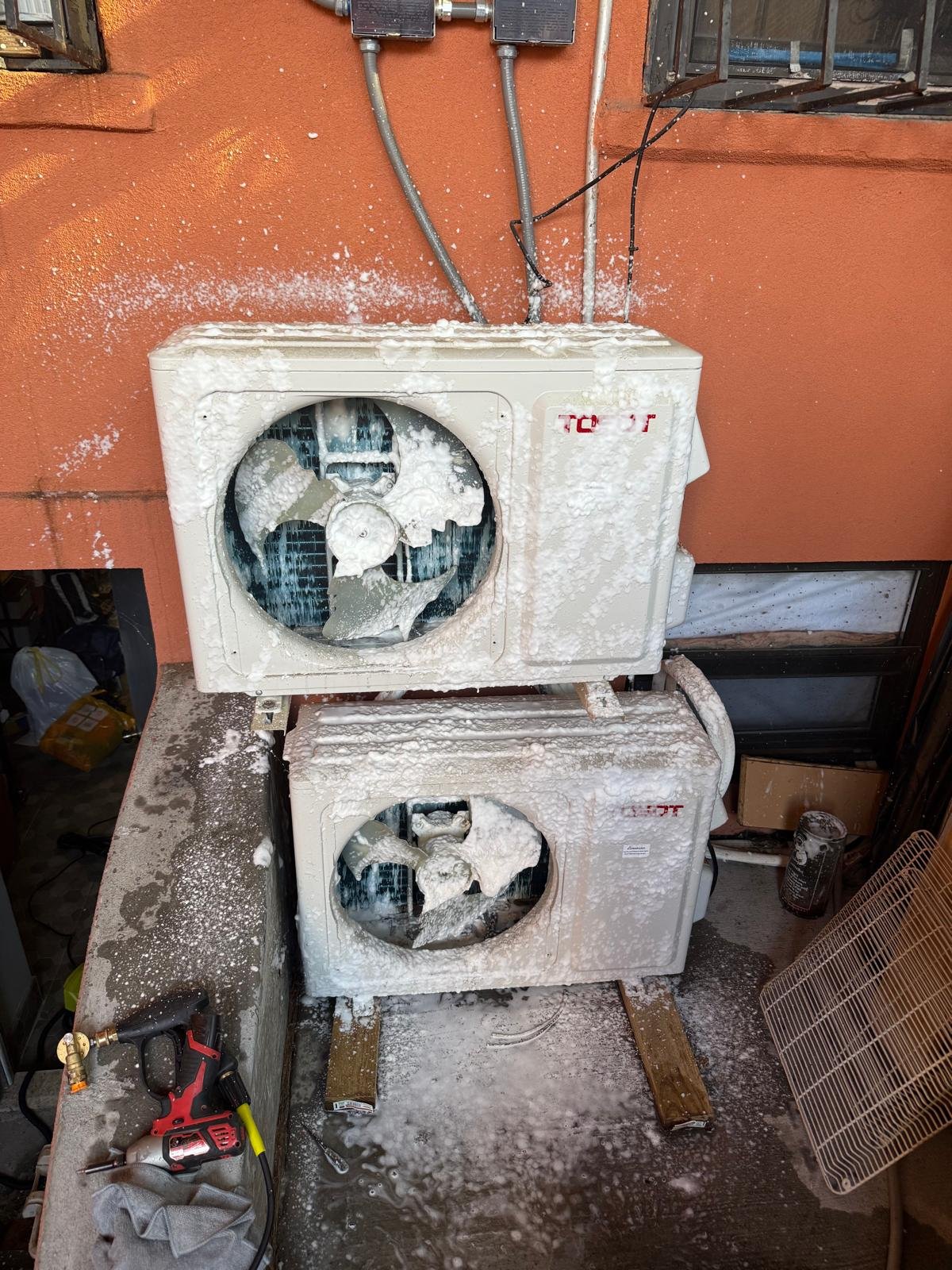 The image shows two outdoor air conditioning units that are covered in large amounts of snow and ice. The units are positioned on a concrete surface next to a reddish-brown wall with electrical wiring and outlets above. Items such as a cordless drill