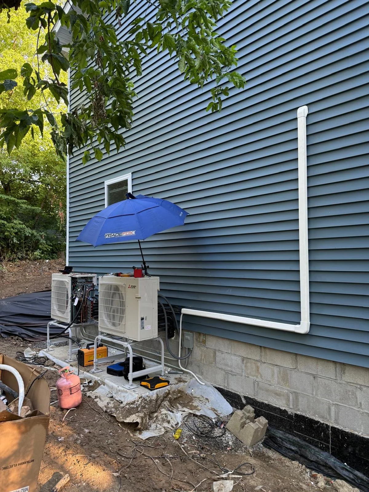 Exterior view of a house wall with air conditioning units and electrical wiring. A blue umbrella is mounted above the units, providing shade. Construction materials and tools are scattered on the ground nearby.