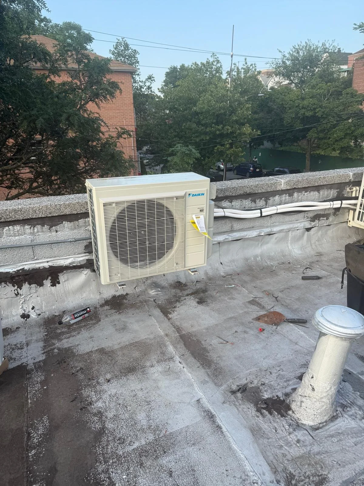 Outdoor rooftop with an air conditioning unit, a plastic bottle on the ground, and various tools and debris. Trees and buildings are visible in the background.