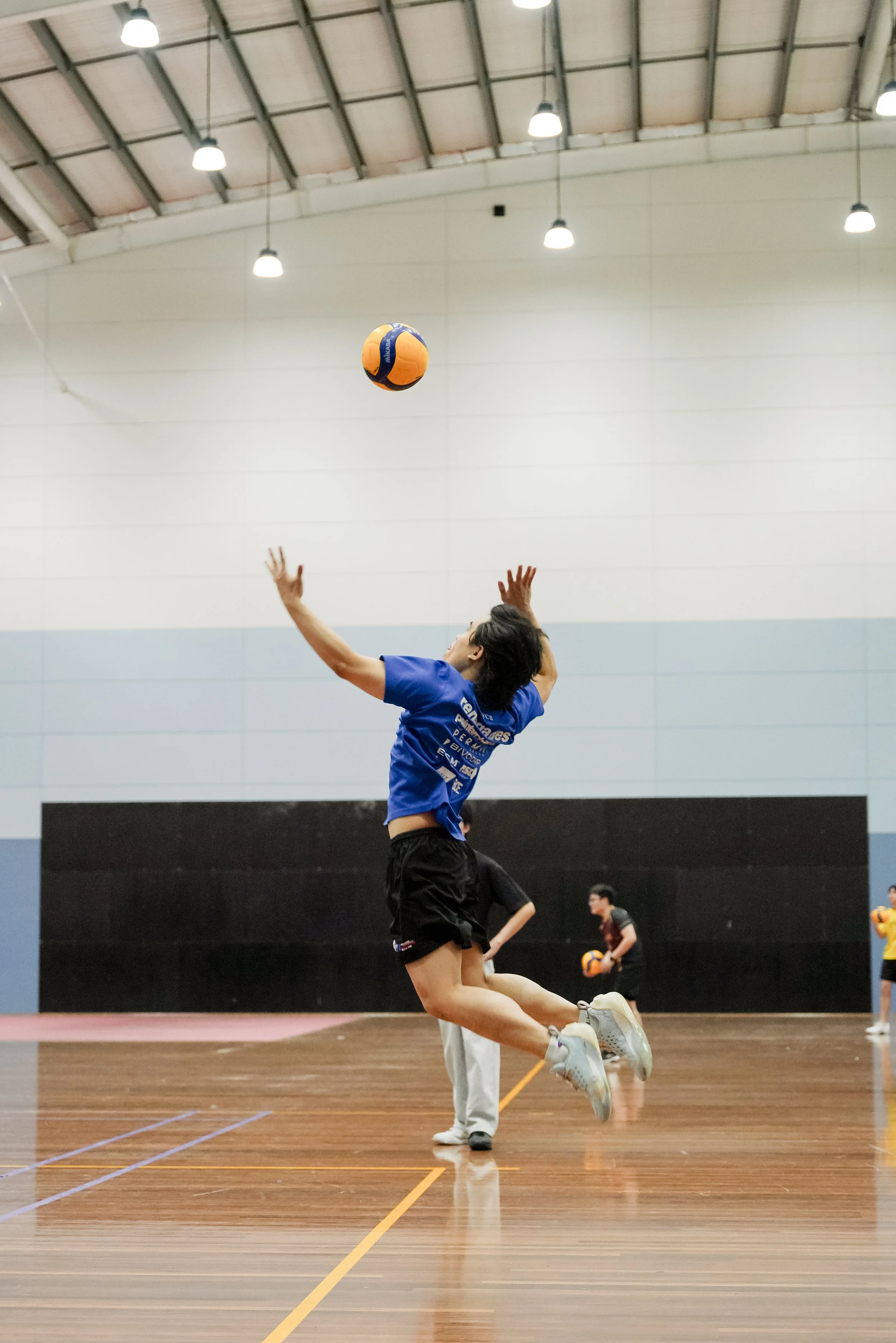 A woman jumping in an indoor volleyball court, preparing to hit a volleyball. Other players are visible in the background.