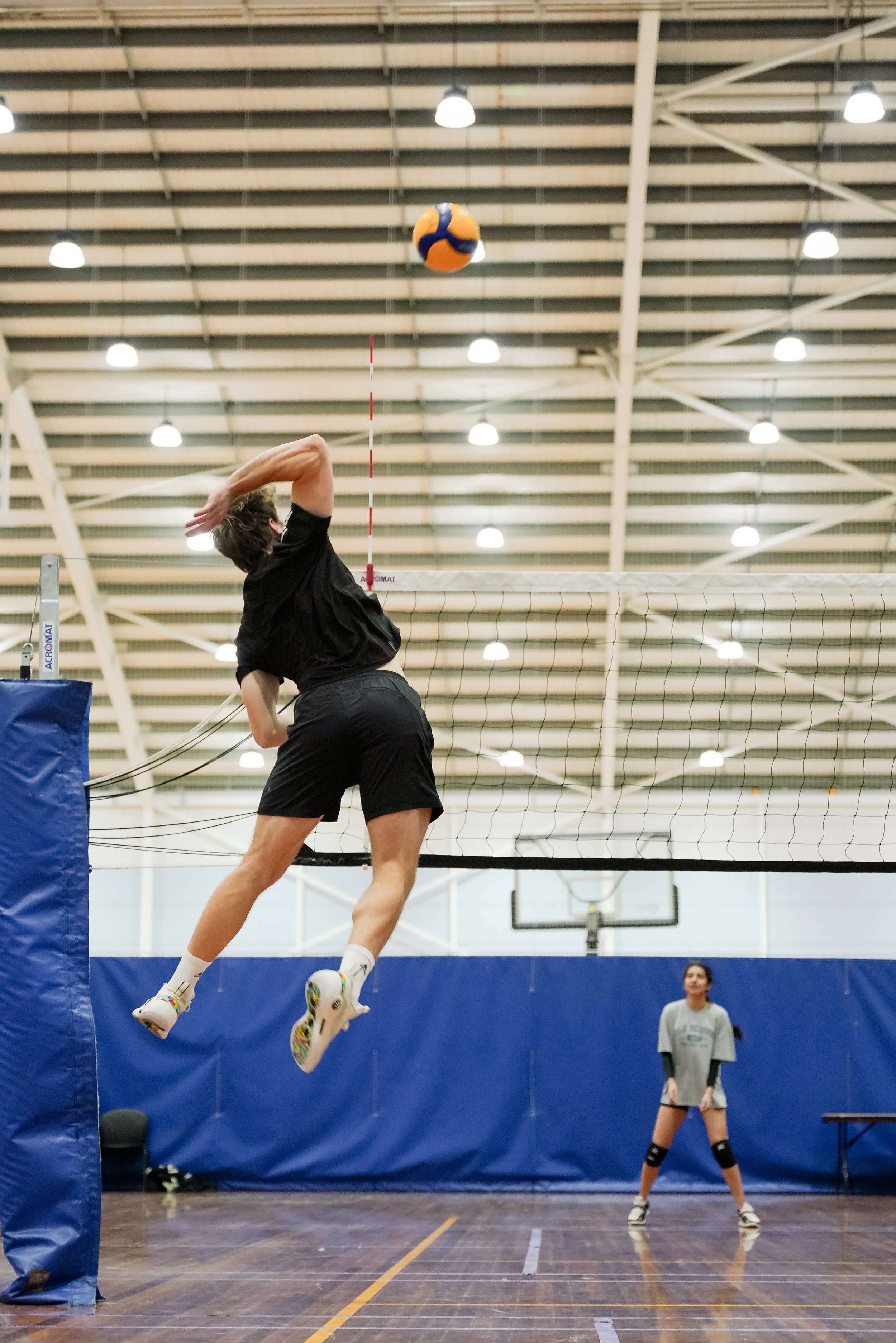 A volleyball player jumps to hit the ball over the net in an indoor gym, with another player standing ready in the background.