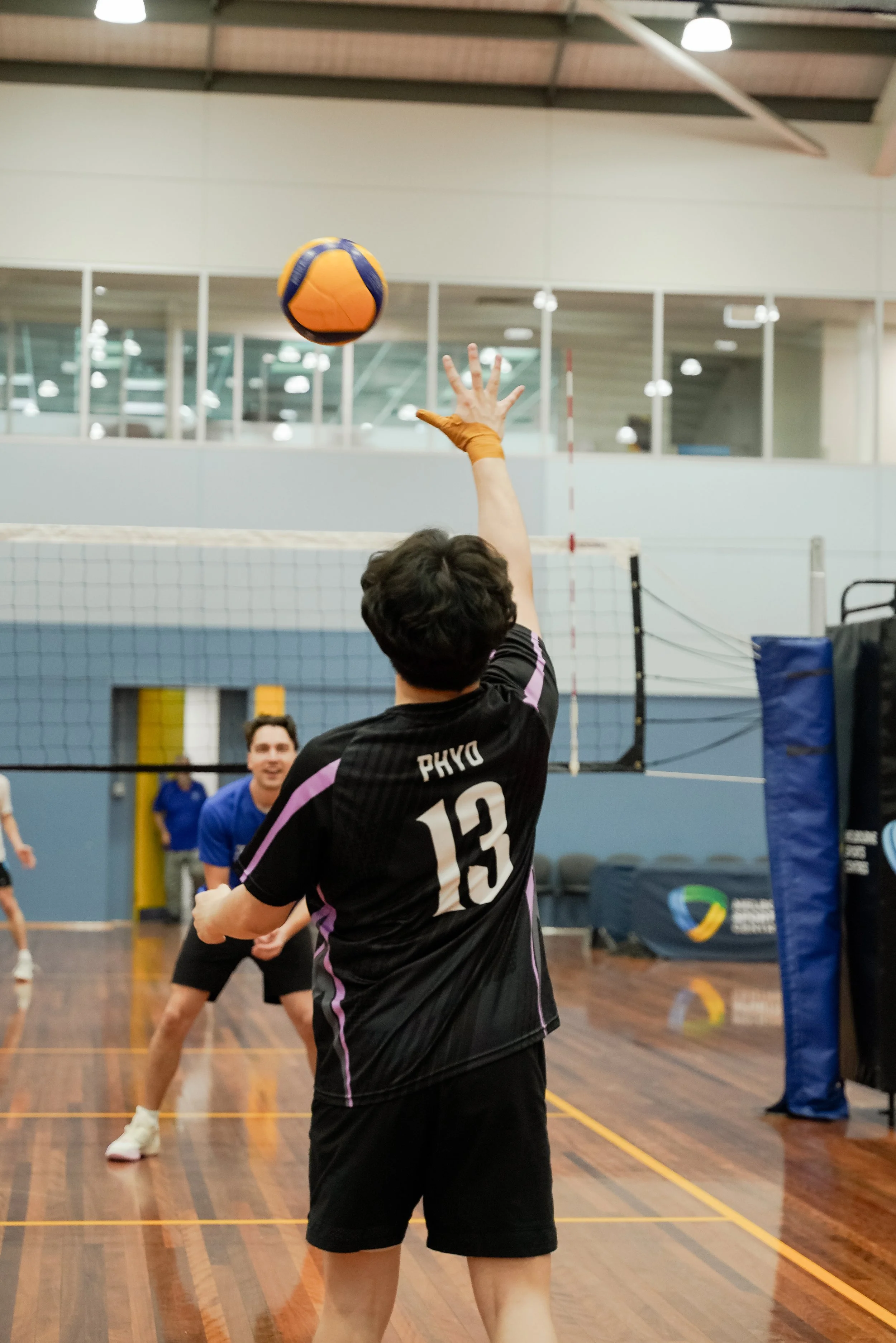 A person with jersey number 13 reaching up with their right hand to hit a volleyball during a game at an indoor gymnasium. There are other players in the background and a volleyball net.
