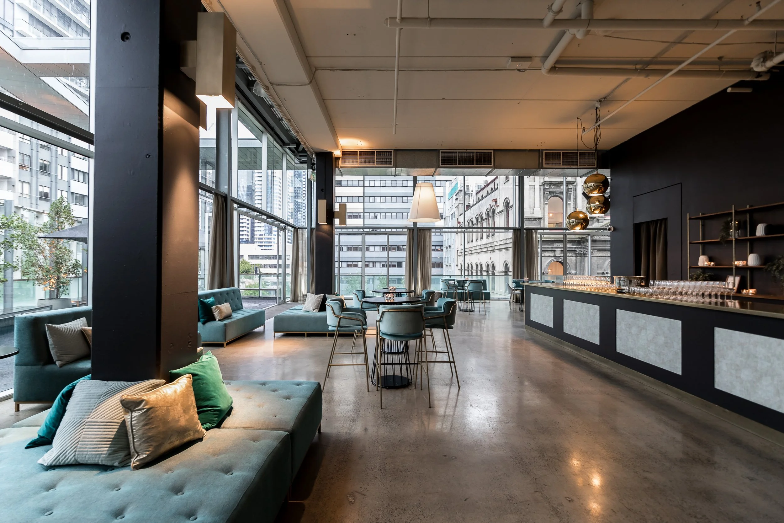 Modern city-view interior lounge with teal sofas and pillows, round tables, large windows, and city buildings outside.