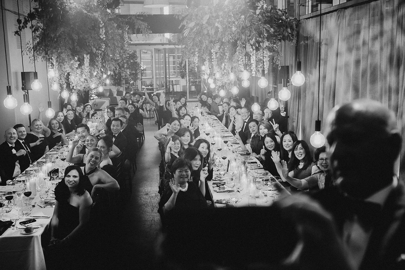 A large group of people sitting at a long dining table, smiling and waving, during a banquet or celebration in a well-lit, modern restaurant or event space.