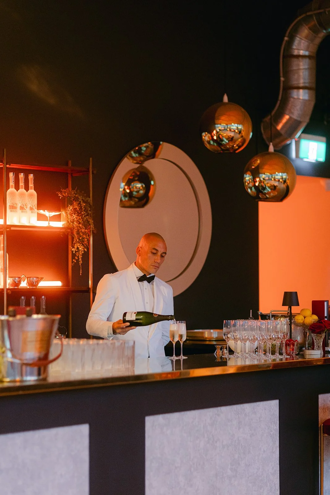 A bartender in a white tuxedo pouring champagne into glasses at a modern bar with warm lighting and decorative spherical pendant lights.