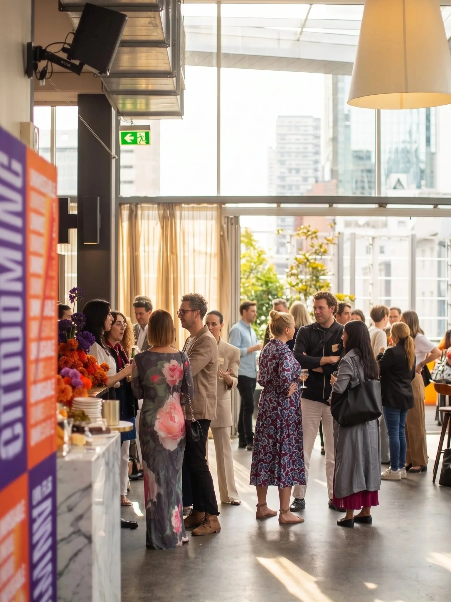 People attending a social event or reception in a large, modern indoor space with floor-to-ceiling windows and cityscape view. Attendees are engaged in conversations, with some holding glasses of wine, and there are floral arrangements and a refreshment table visible.