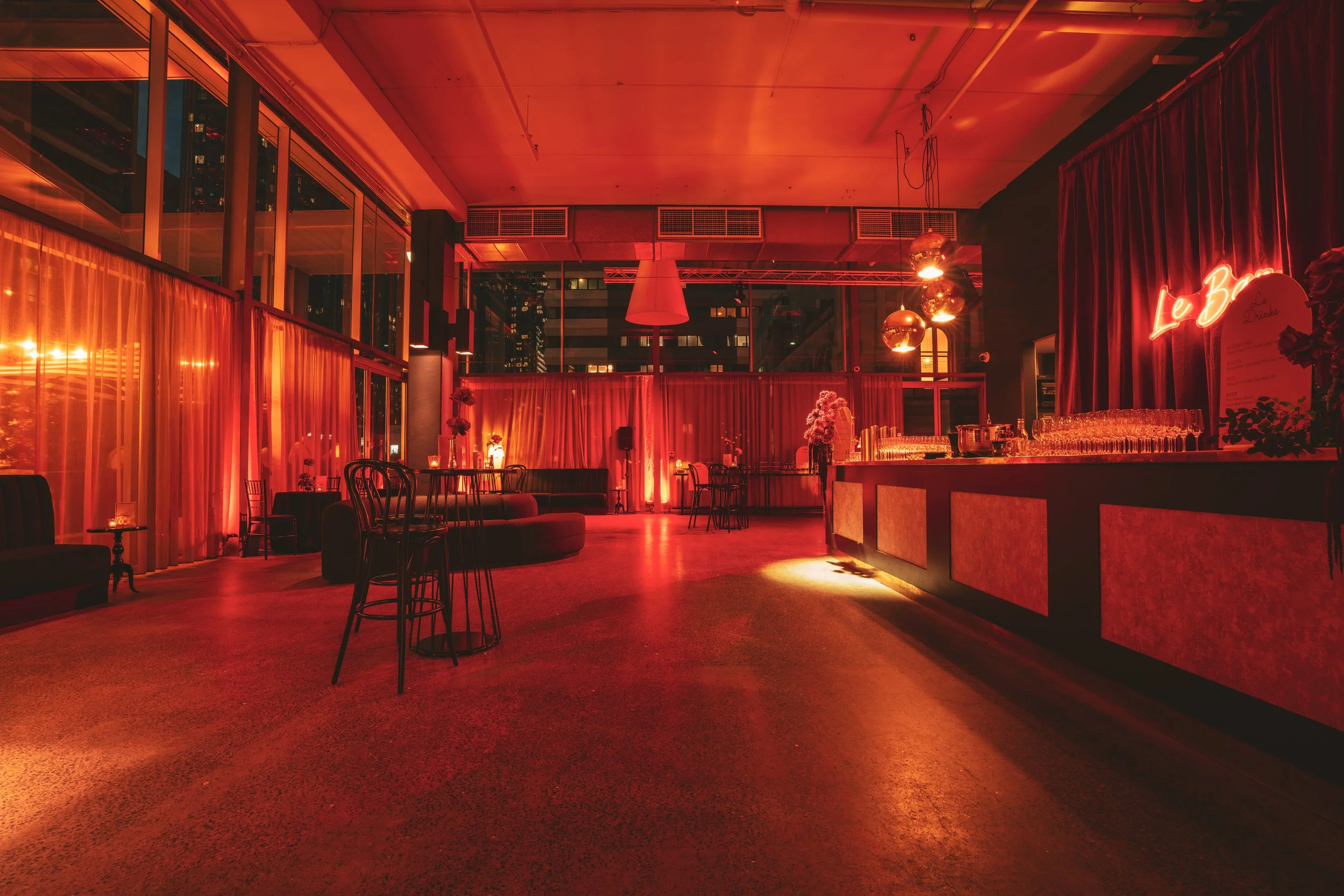 Empty lounge or bar area illuminated with red lighting, featuring tables, chairs, a bar counter with glasses, and decorative flowers, with large windows and city lights outside.
