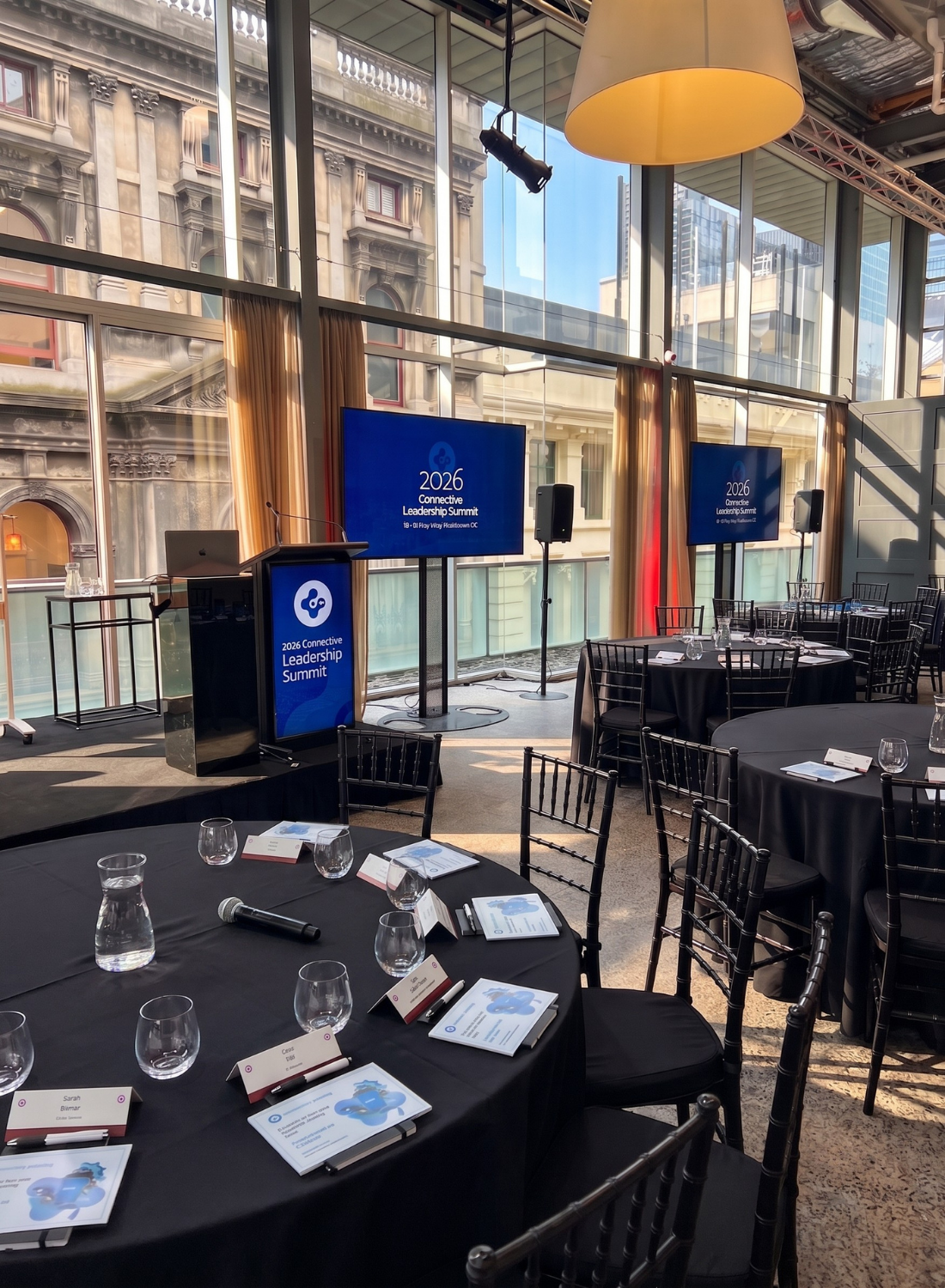 Conference room set up for the 2026 Connective Leadership Summit, featuring round tables with black tablecloths, place cards, water glasses, and brochures. There are large screens displaying the event logo, a podium with a microphone, and a window view of nearby historic buildings.