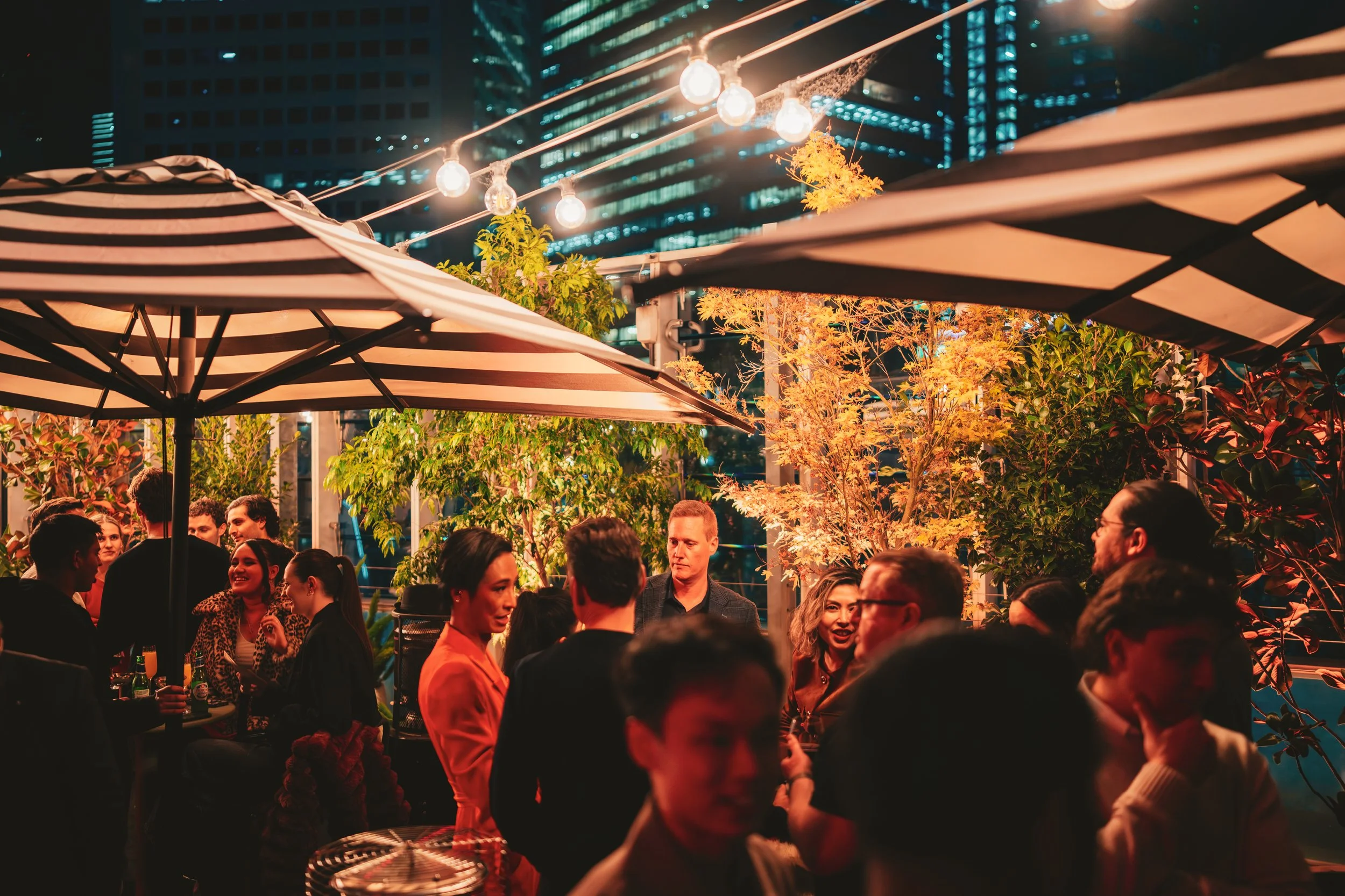 Nighttime outdoor gathering with people talking, surrounded by lush greenery, striped umbrellas, string lights, and city skyscrapers in the background.