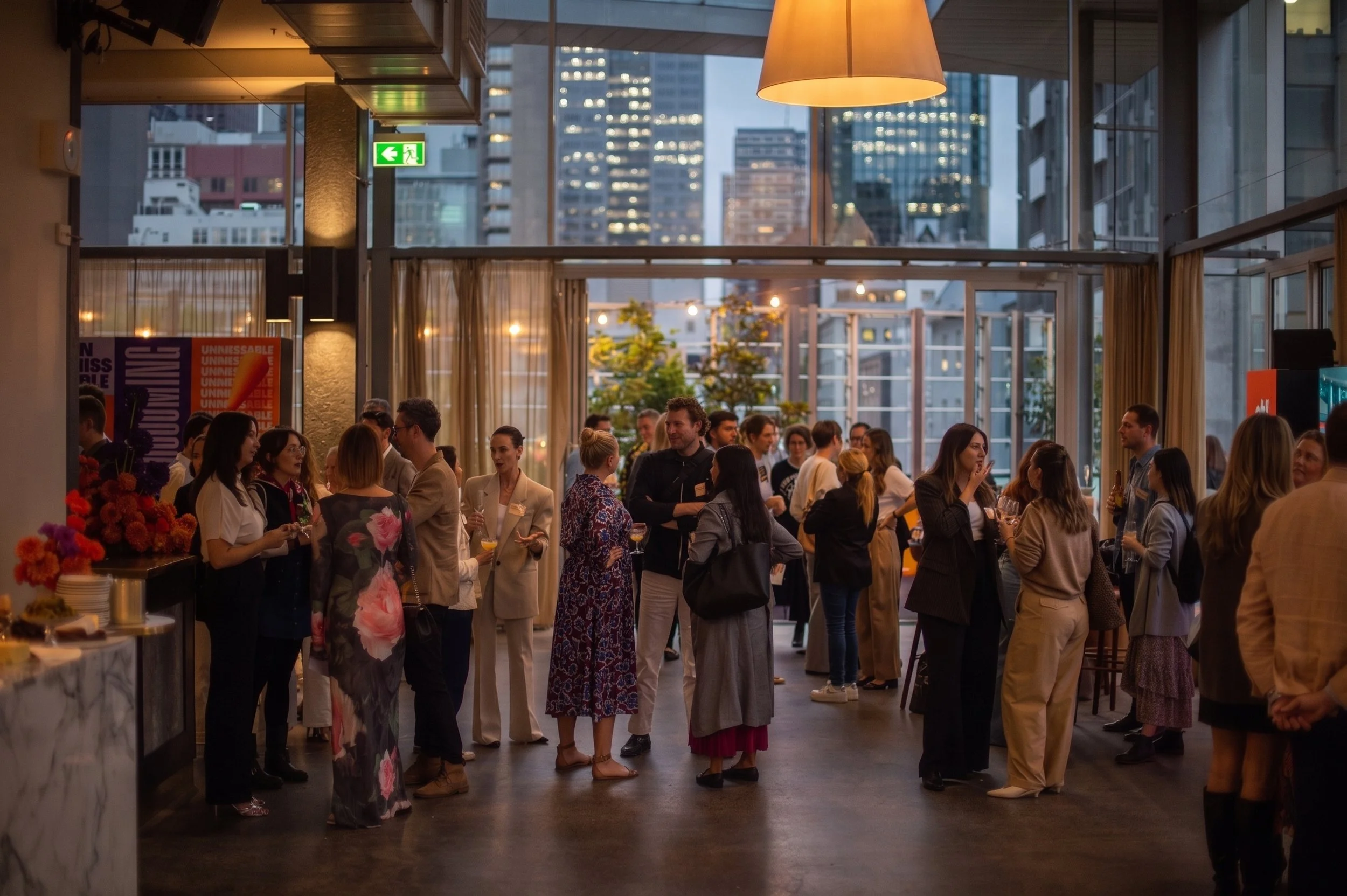 People socializing at an indoor event in a cityscape setting during evening with tall buildings and city lights visible through large windows.