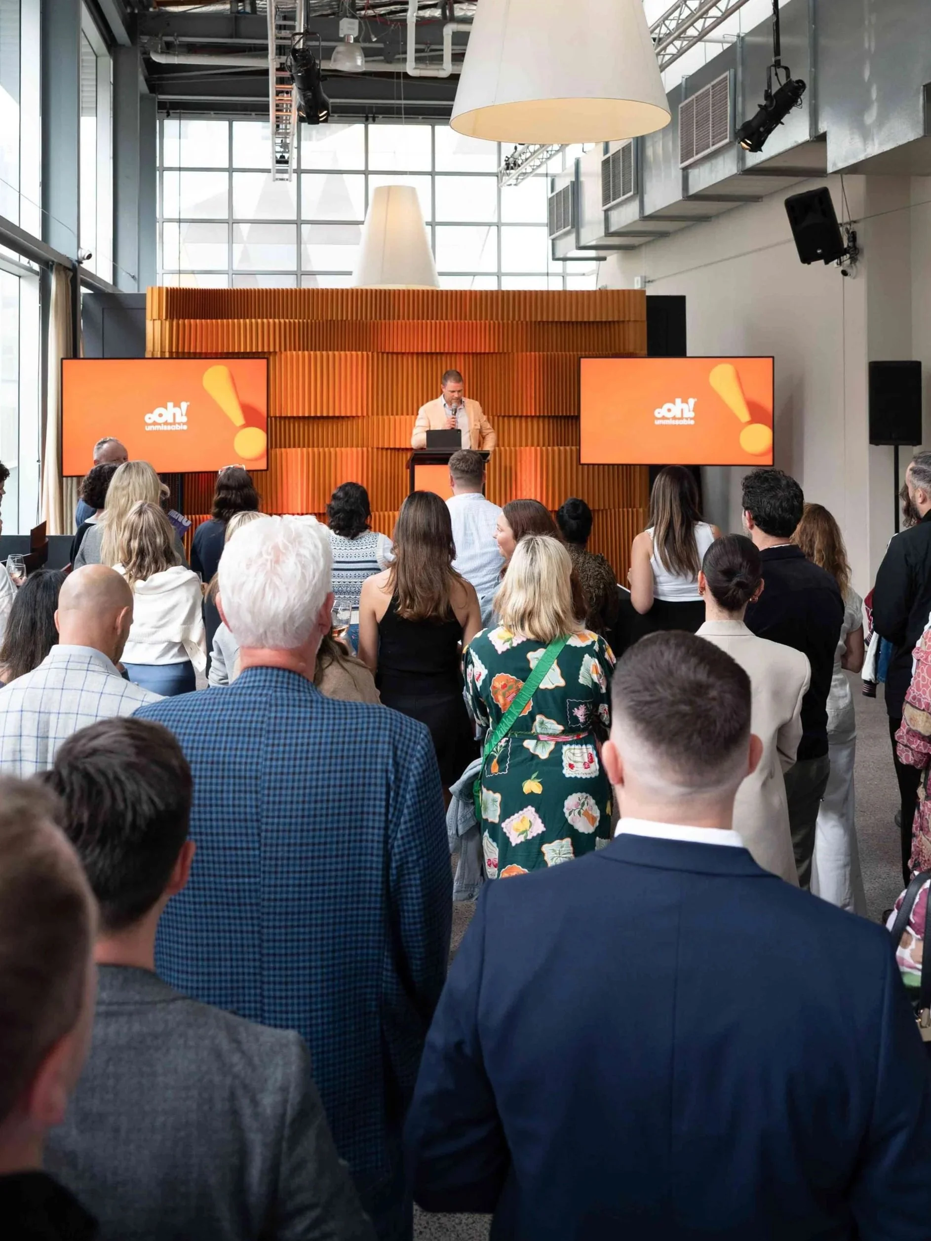 An indoor conference room with a speaker at a podium, speaking to an audience. The room has large windows, modern decor, and two large screens displaying the "OAH! Unmistakable" logo.