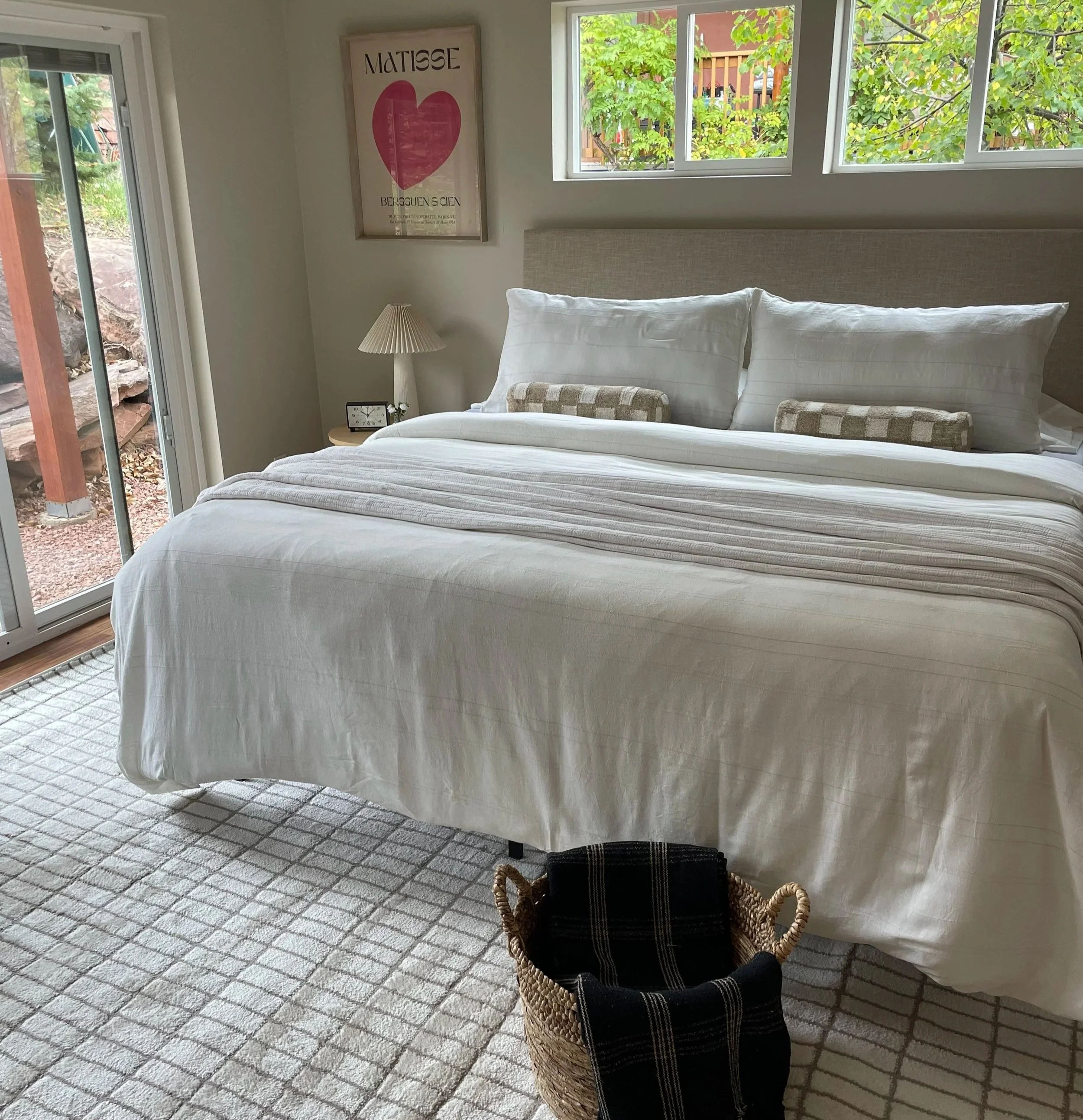 A bedroom with a hamper in the foreground and a crisp white bed behind it with a Matisse poster in the back.