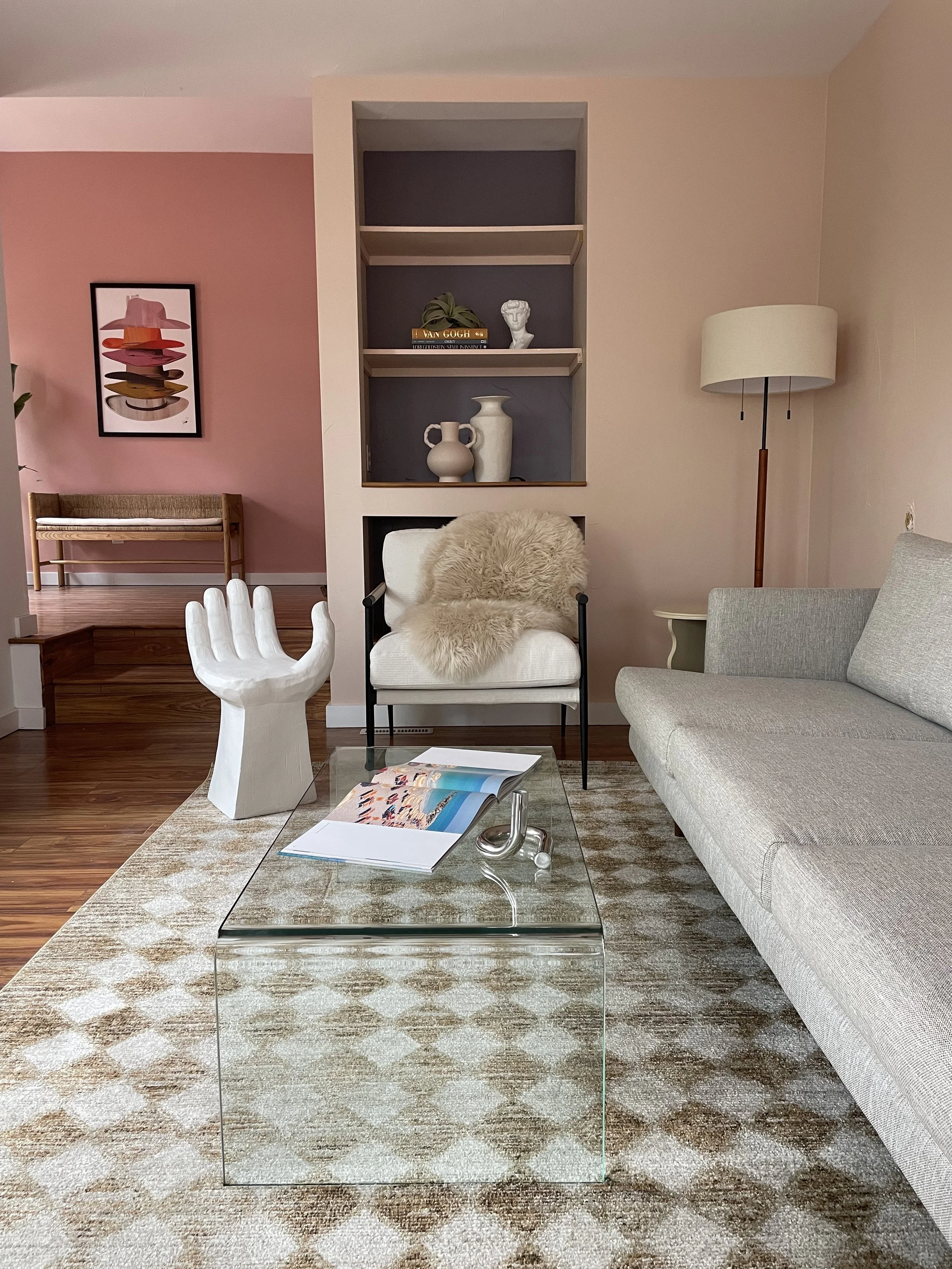 A light pink living room with a couch, glass table, stool in the shape of a hand, and a lounge chair with a fur blanket on top.