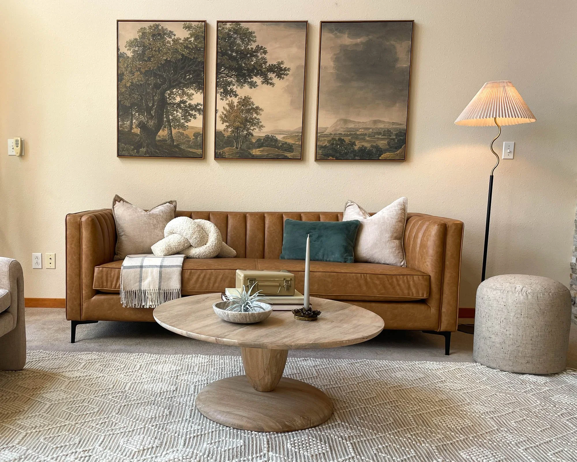 A living room with a caramel brown couch, a marble round coffee table, and 3-paneled landscape artwork in the background.