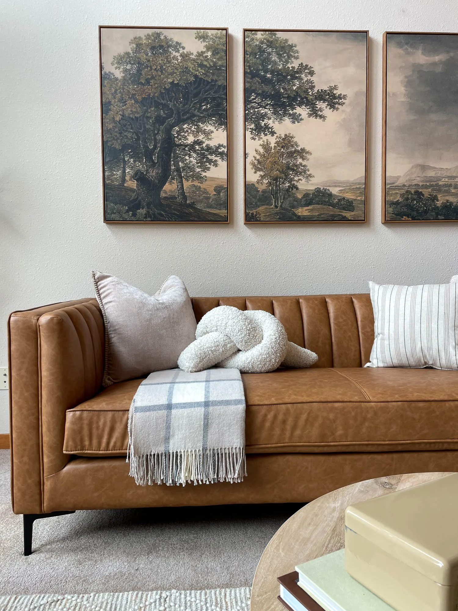 A caramel leather couch with blankets and 3 paneled landscape artwork behind it.