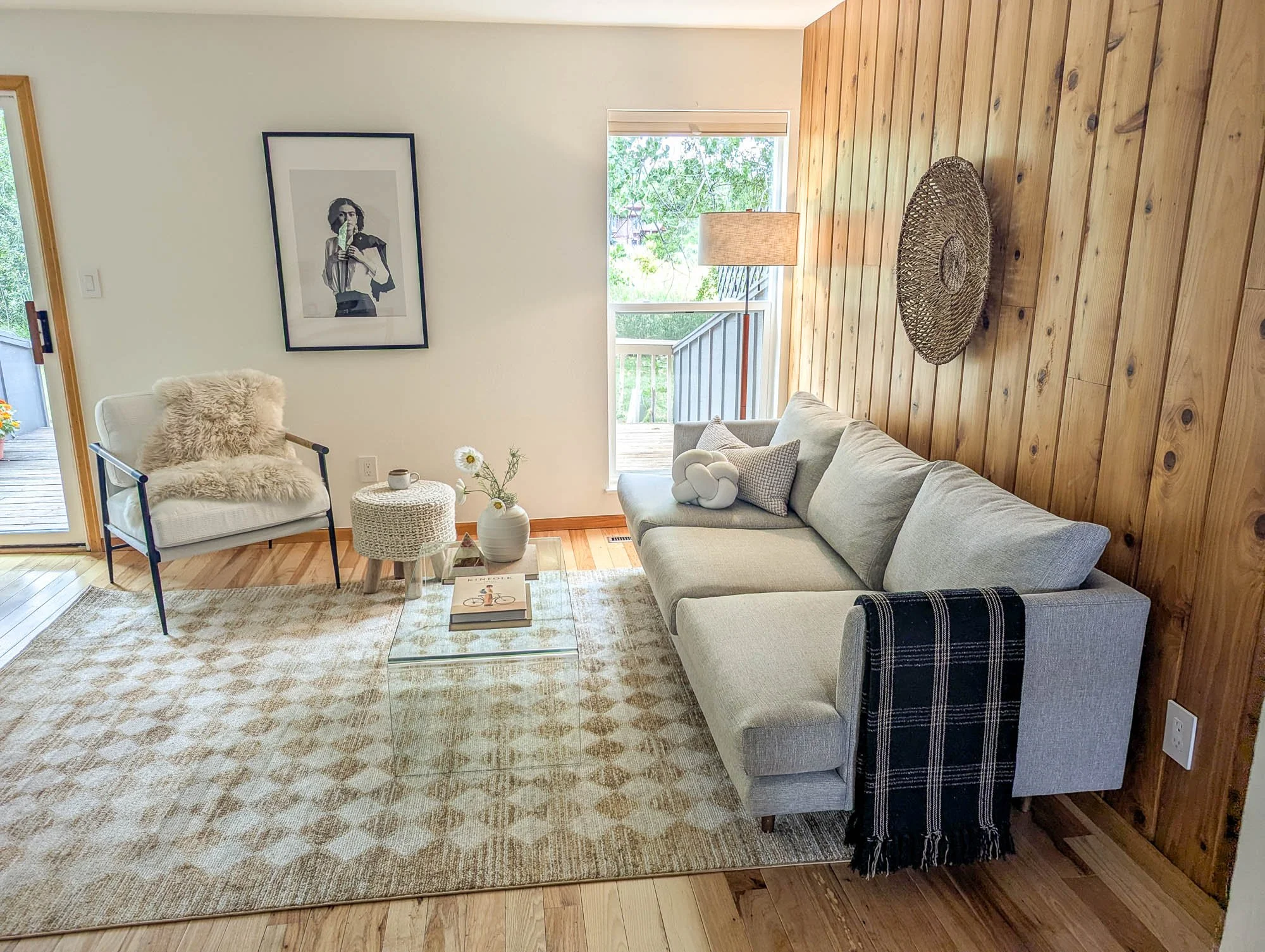 A living room with a photo Frida Kahlo, a grey couch, a lounge chair with a fur blanket, and a glass table.