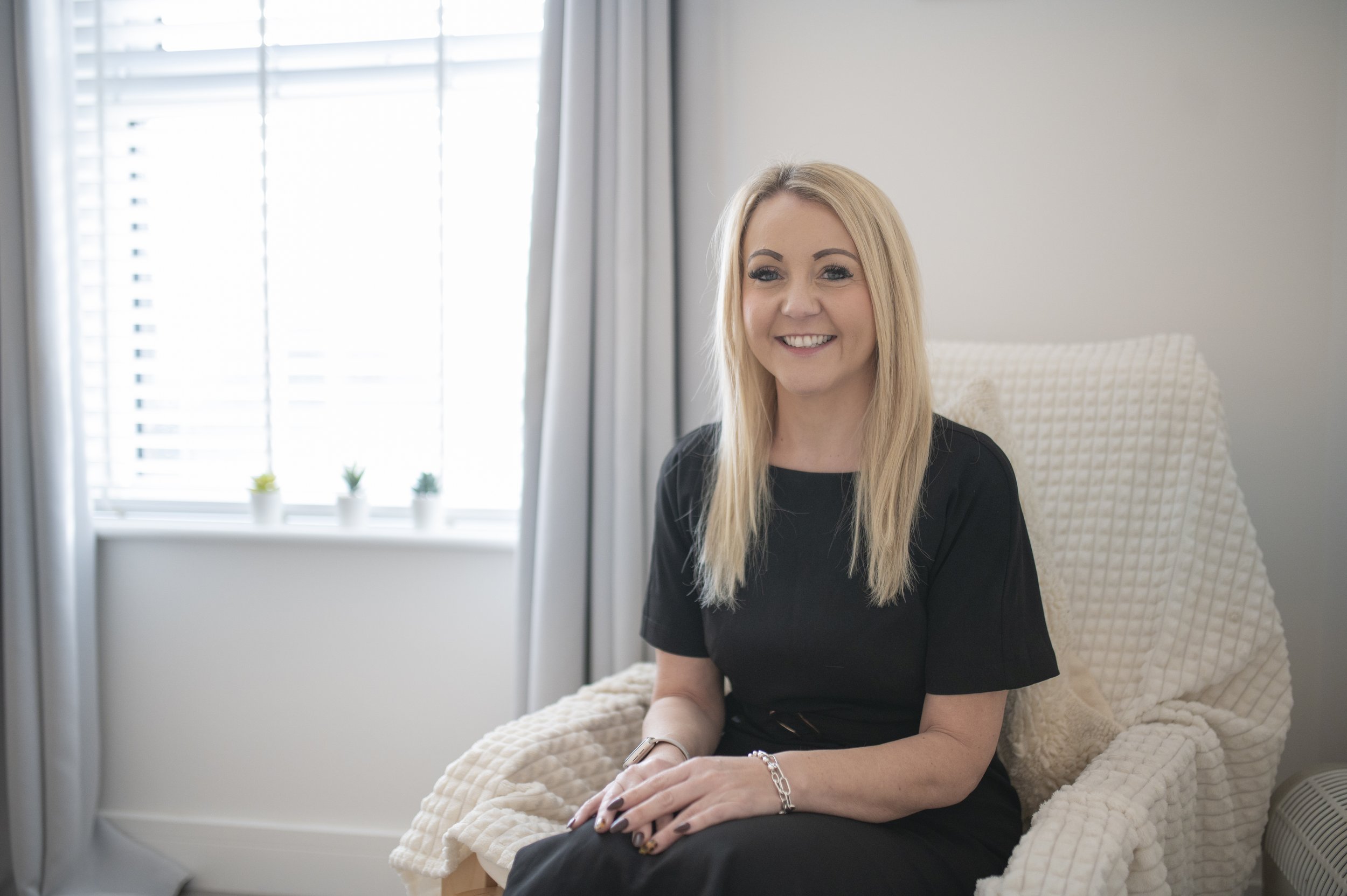 A blonde woman wearing a black shirt sitting on a beige armchair in a well-lit room with white walls, a window with blinds, and small potted plants on the windowsill.