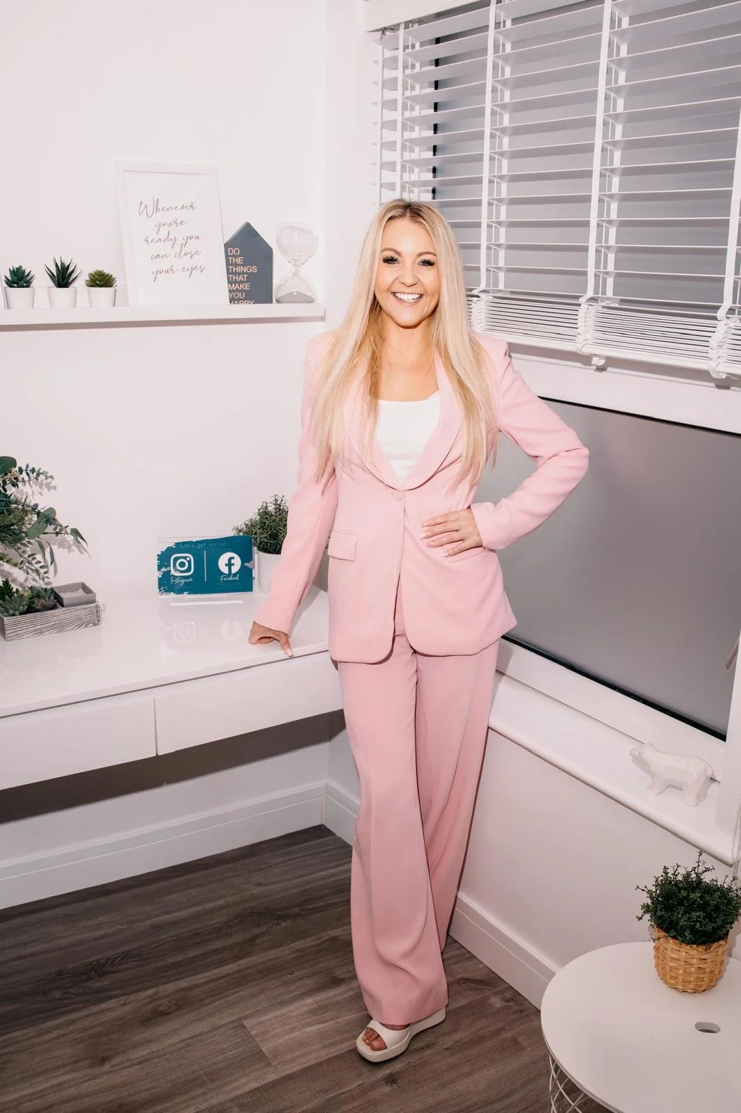 A woman in a pink suit smiling, standing in a modern white room with plants, signs, and a large television.