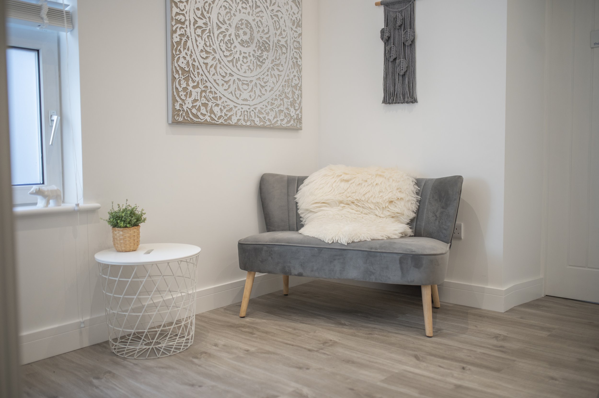 A cozy corner with a gray upholstered armchair accented with a fluffy cream-colored throw pillow and a matching throw blanket. To the left, there's a small white side table with a woven basket holding a green plant. On the window sill, there's a small decorative horse figurine. The wall features a large, intricate beige carving and a gray woven wall hanging. The room has light-colored laminate flooring and white walls.