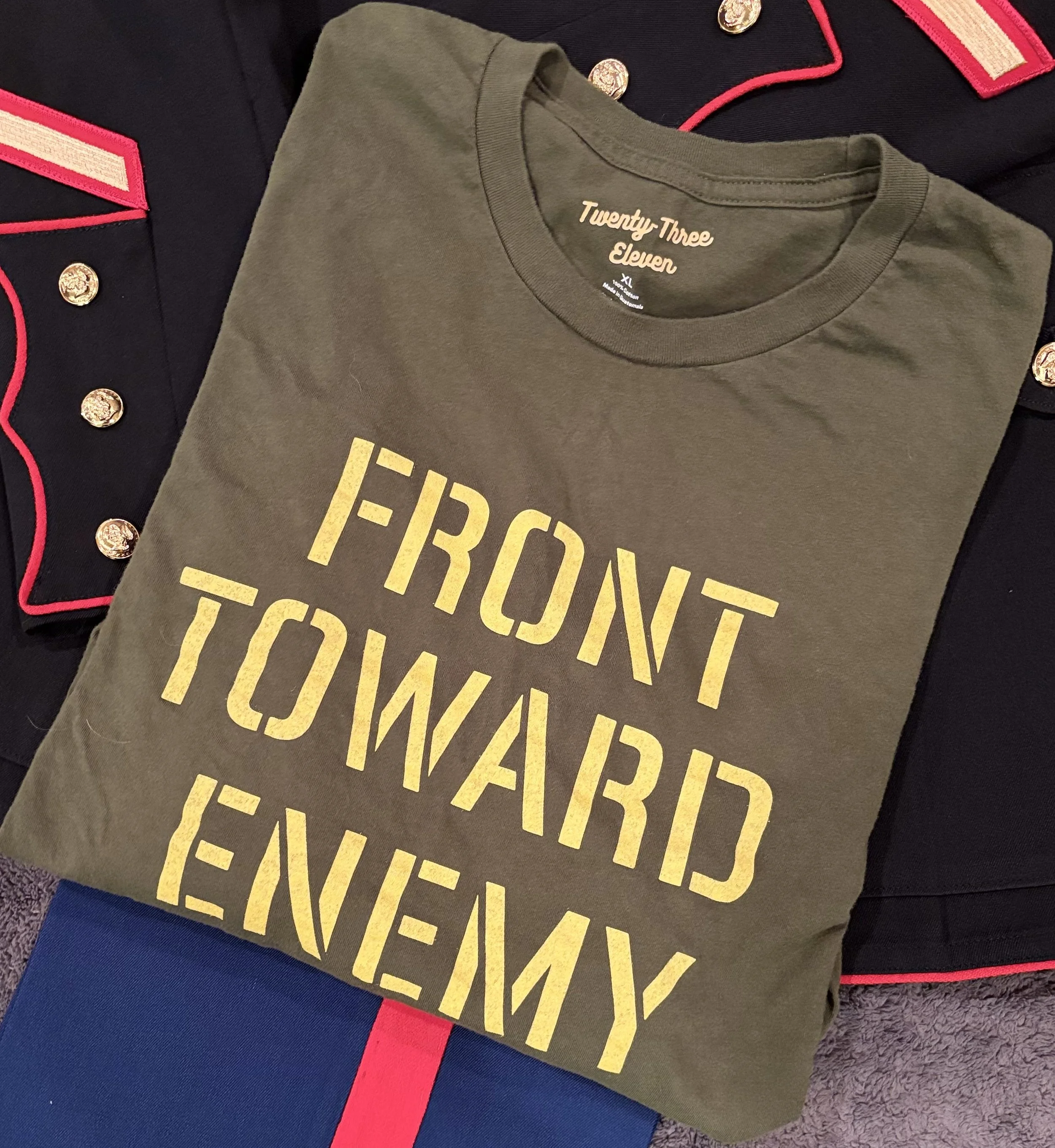 A folded green T-shirt with yellow text reading 'FRONT TOWARD ENEMY' lies on top of a U.S. Marine Corps dress blue uniform.