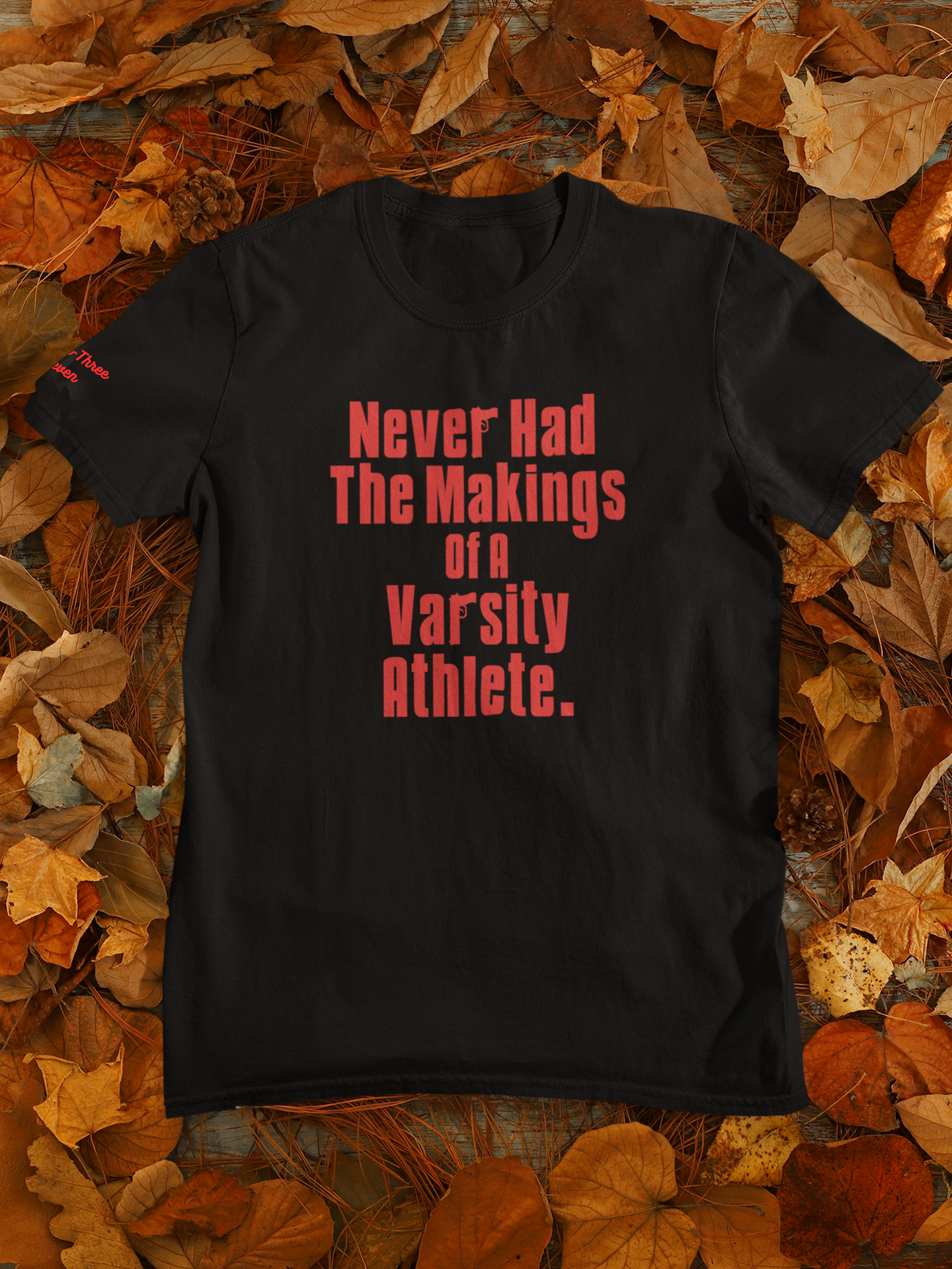 Black T-shirt with red text that reads "Never Had The Makings of a Varsity Athlete" lied on a surface covered with autumn leaves.