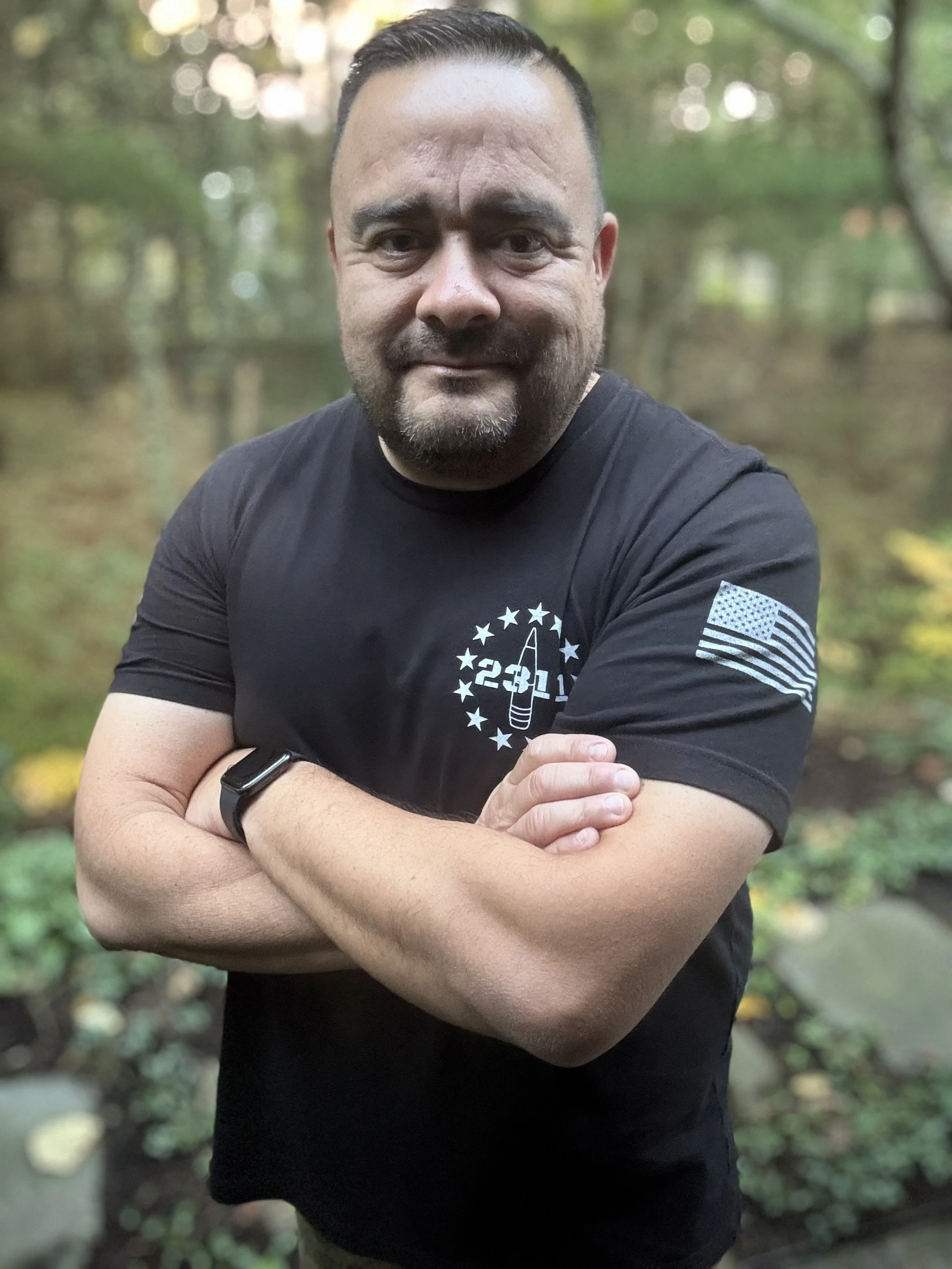 A man with short dark hair and a beard, wearing a black T-shirt with a graphic and American flag patch on the sleeve, standing outdoors with trees and greenery in the background, arms crossed, smiling at the camera.