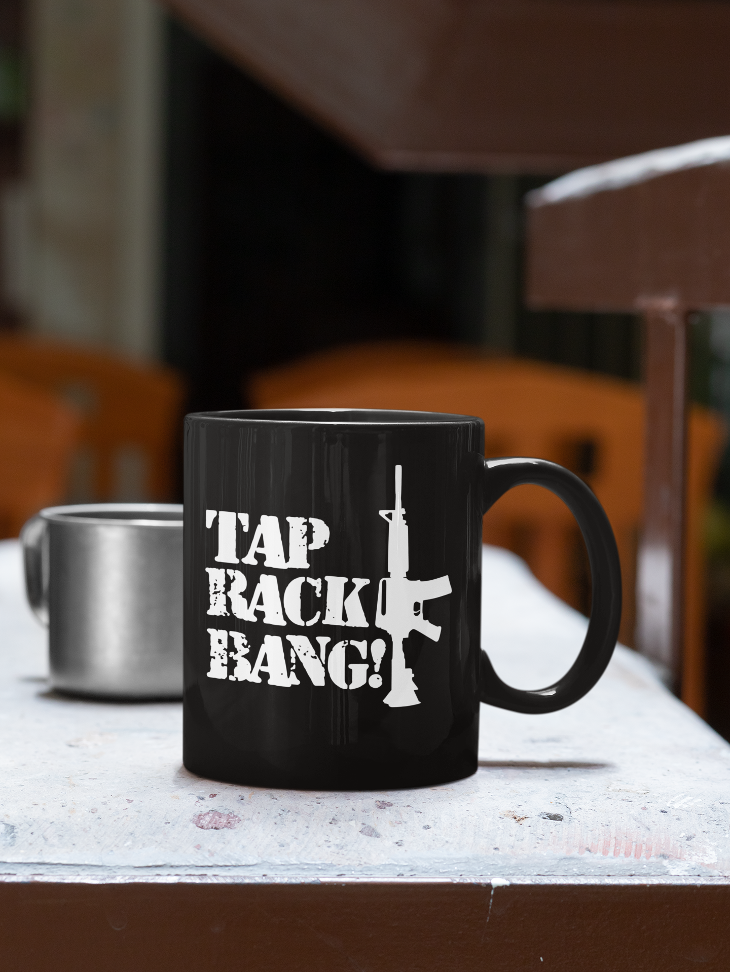 Black coffee mug with white text that says "TAP RACK BANG!" and a white silhouette of a rifle. The mug is on a white surface with a blurred wooden chair and table in the background.