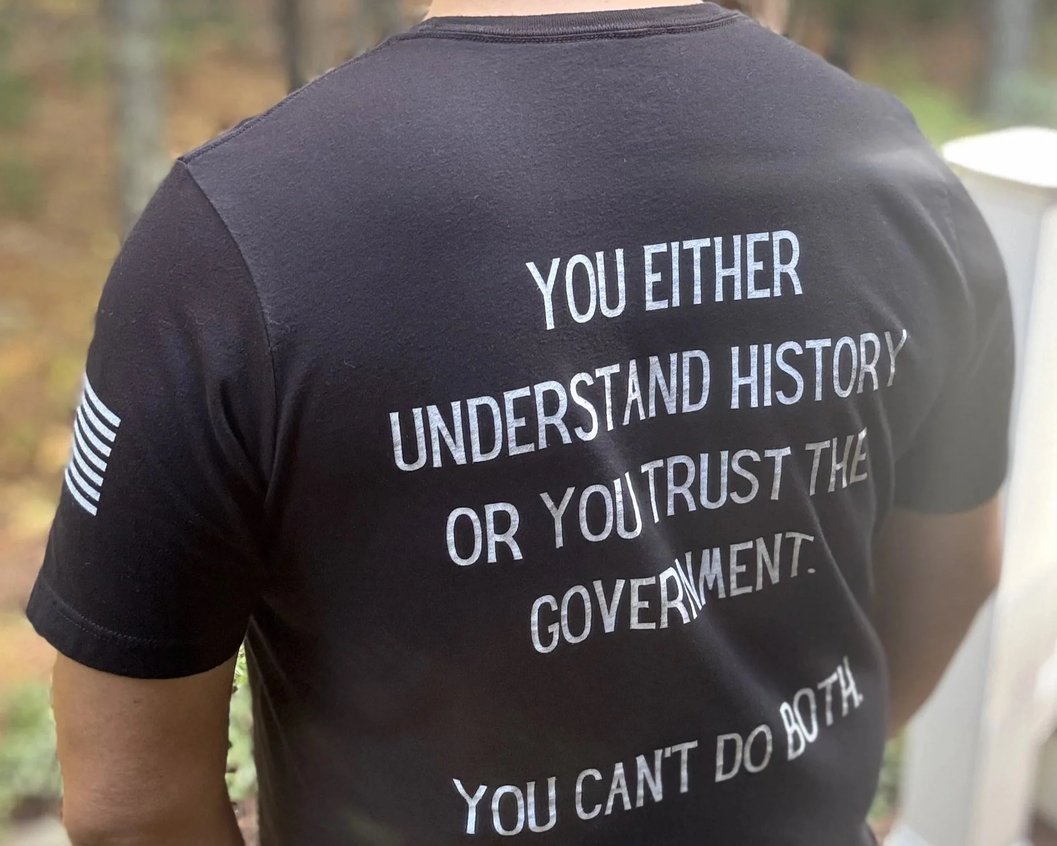 The back of a person wearing a black t-shirt with a quote about understanding history and trusting government, with an American flag patch on the sleeve, outdoors with blurred trees in the background.