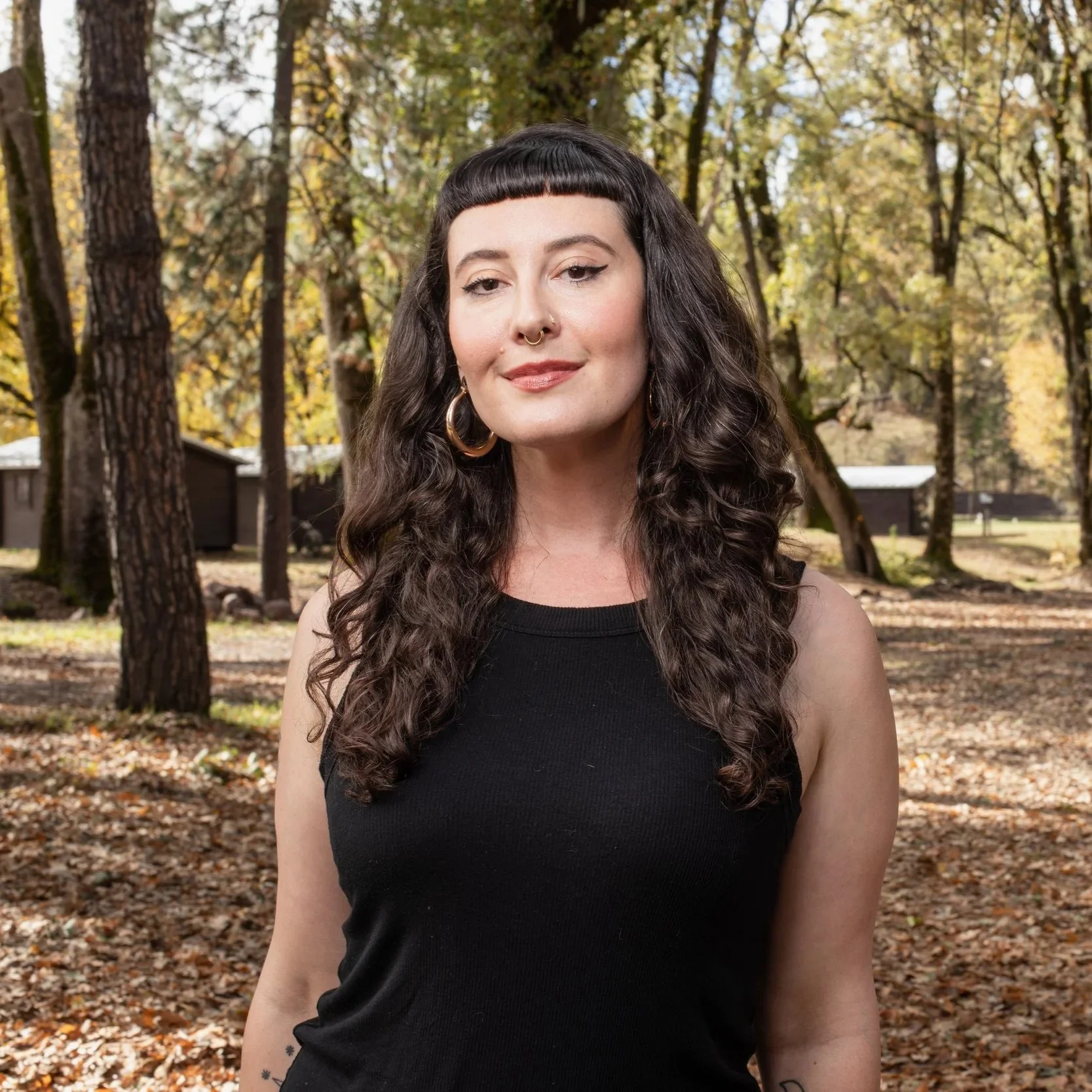 A woman with long, curly dark hair and a nose piercing, standing in a forested area with trees and fallen autumn leaves.