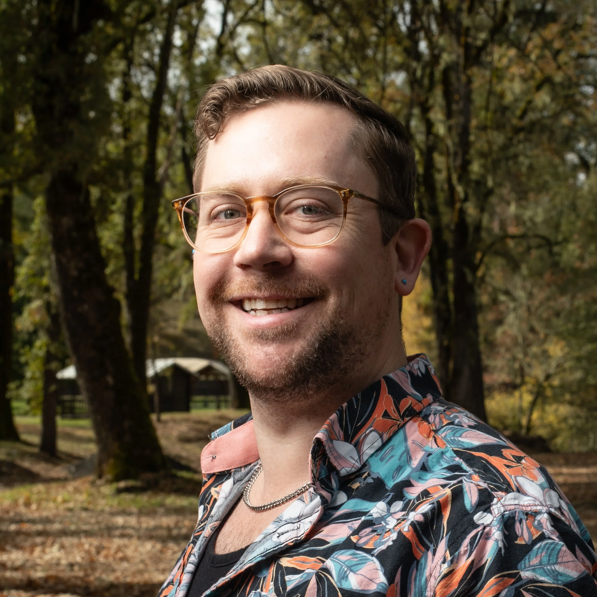 A smiling man with glasses and a colorful patterned shirt standing outdoors in a wooded area with trees in the background.