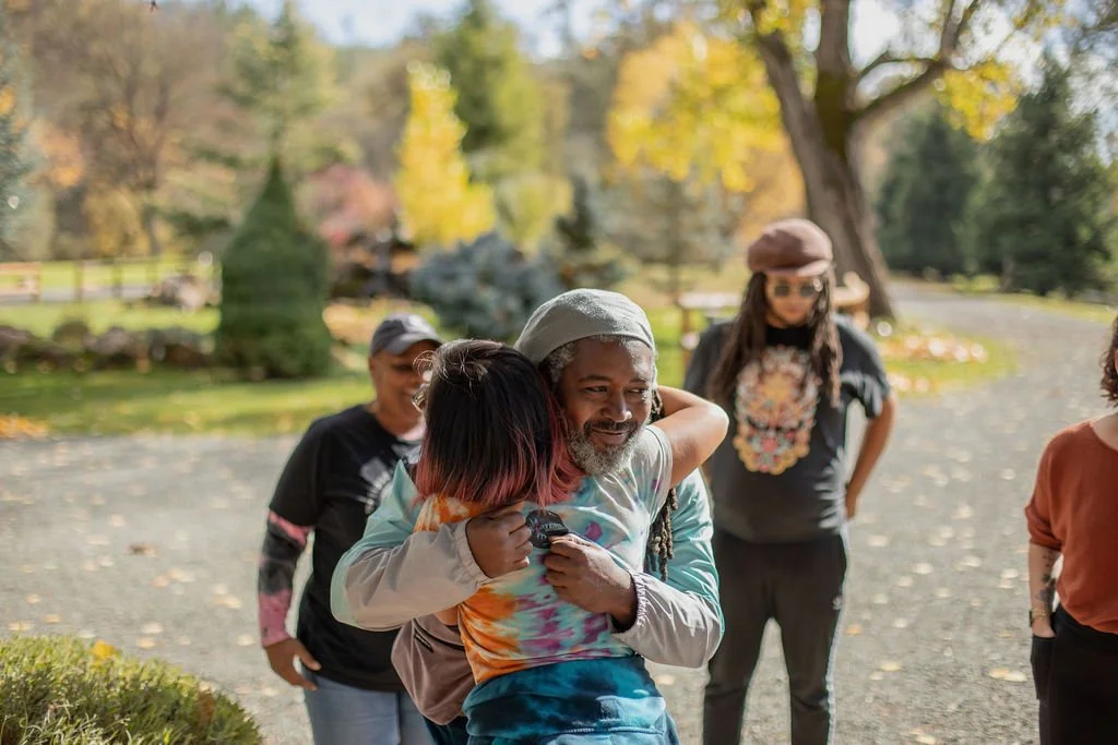 Group hug among four friends outdoors in a park during autumn, with colorful fall trees and a gravel path in the background.