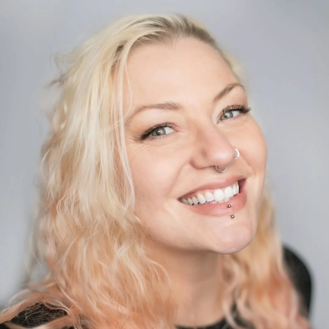 Close-up of a smiling woman with blonde, wavy hair, blue eyes, and multiple facial piercings, against a plain background.