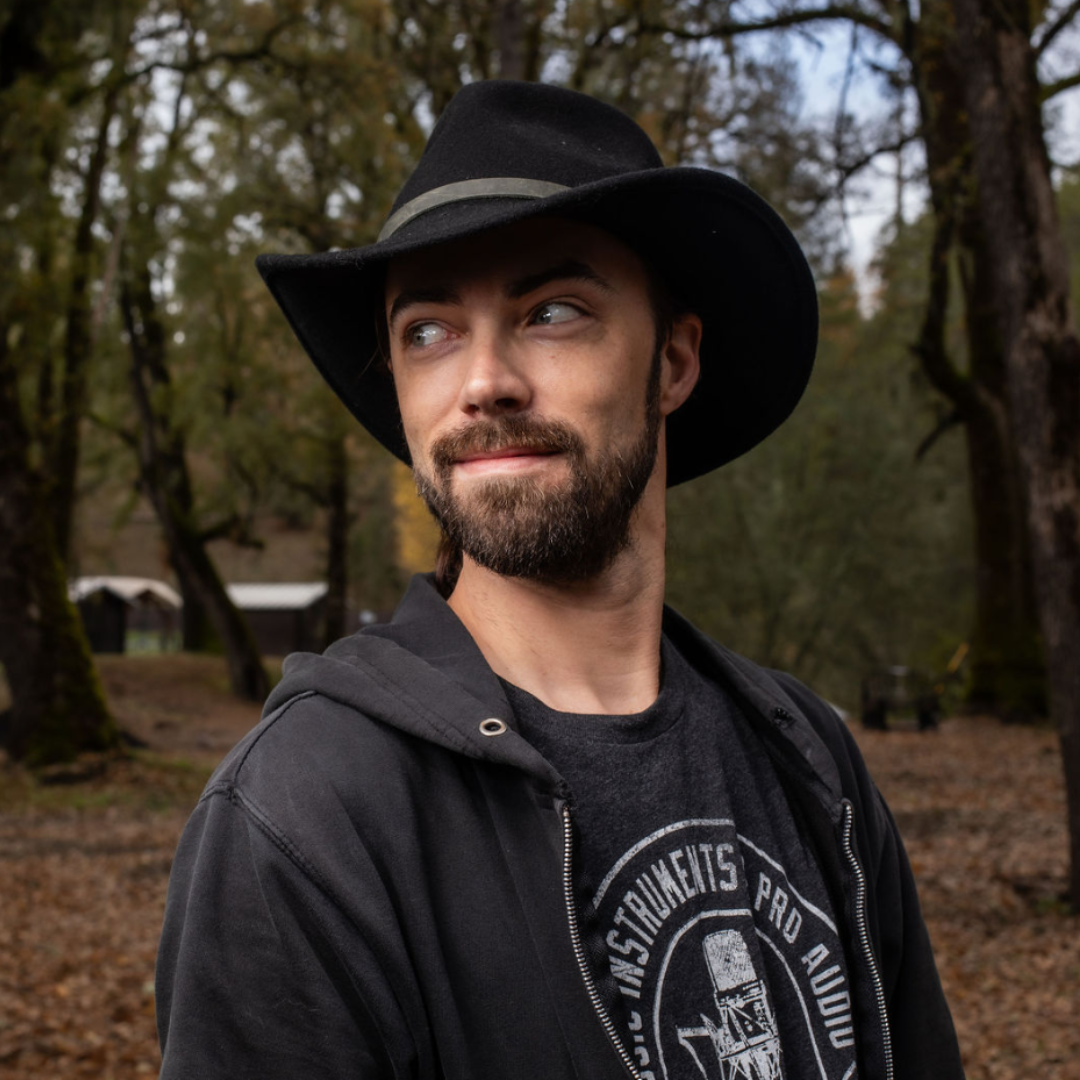 A man with a beard and mustache wearing a black wide-brimmed hat, a black hoodie, and a graphic t-shirt, standing outdoors in a wooded area during autumn.