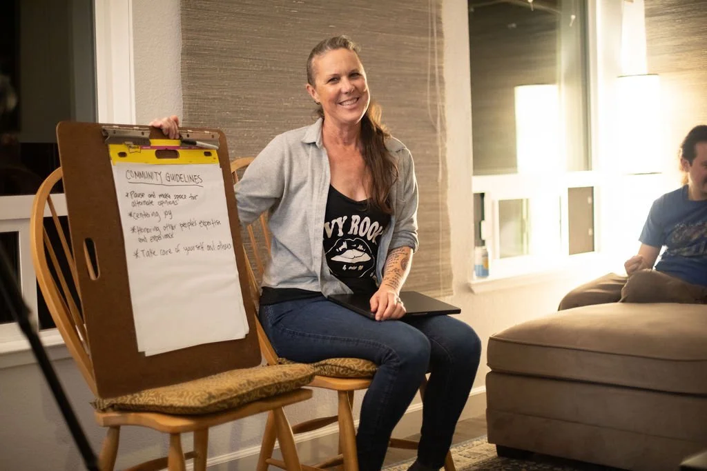 A woman sitting on a chair next to a table with a large pad of paper listing community guidelines. She is smiling, wearing a black t-shirt with a graphic, and has long hair tied back. Two people are sitting on a couch nearby in a warmly lit room with windows and a lamp.