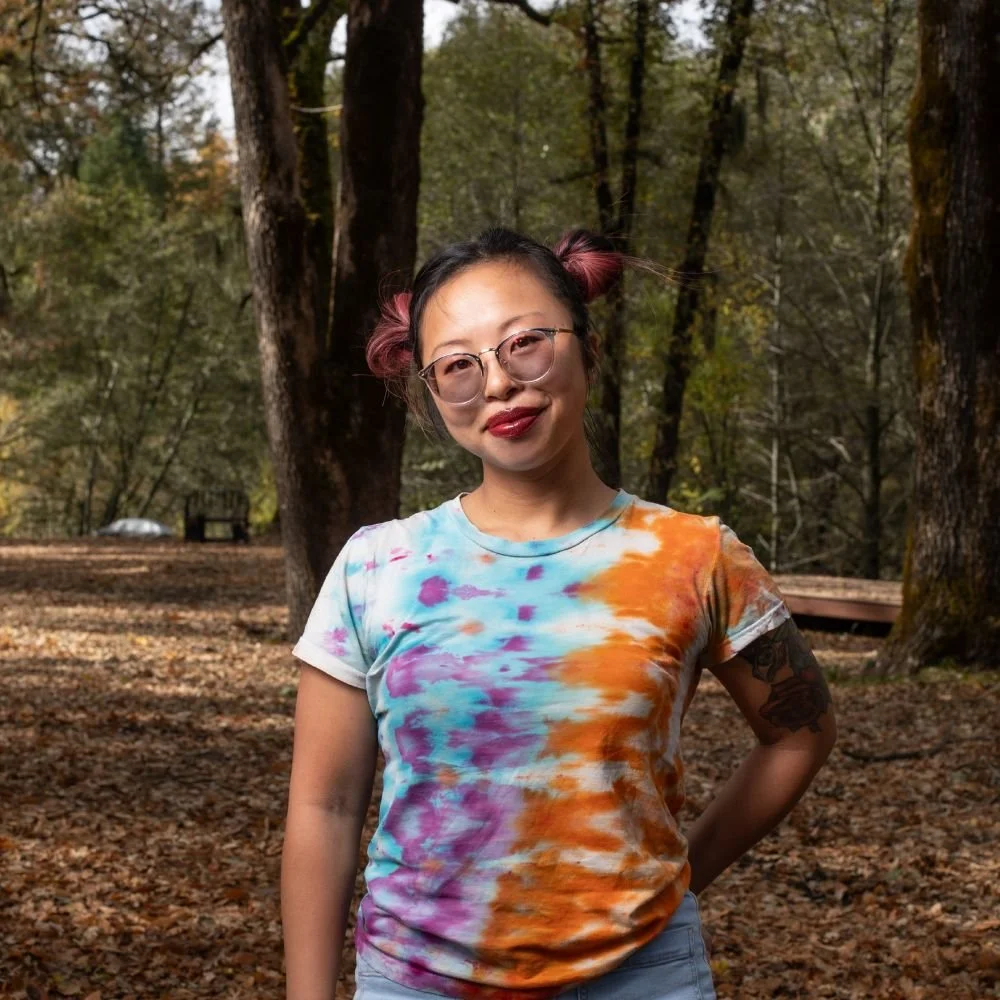 A young woman with glasses and pink hair tied in buns stands in a forest during autumn, wearing a tie-dye T-shirt with vibrant colors.