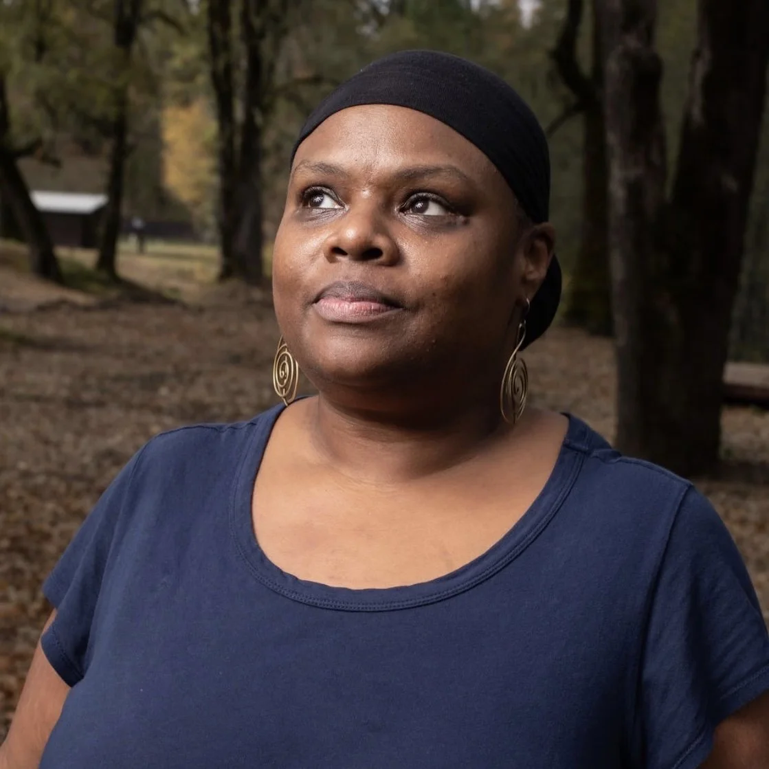 A woman wearing a black headscarf and large spiral earrings outdoors in a wooded area, looking off to the side.