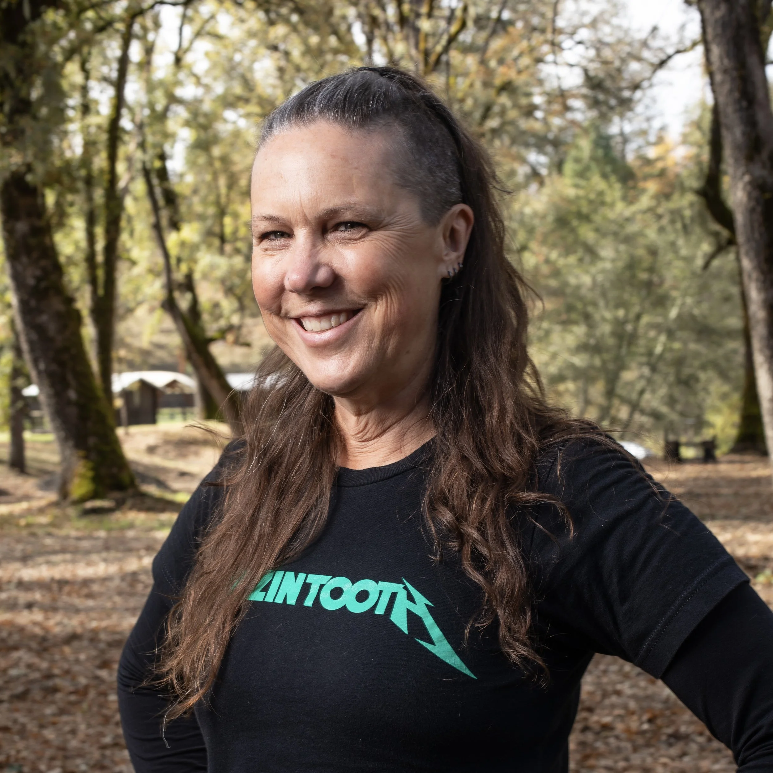 A woman with long brown hair, wearing a black T-shirt with teal text, smiling outdoors in a wooded area during autumn.