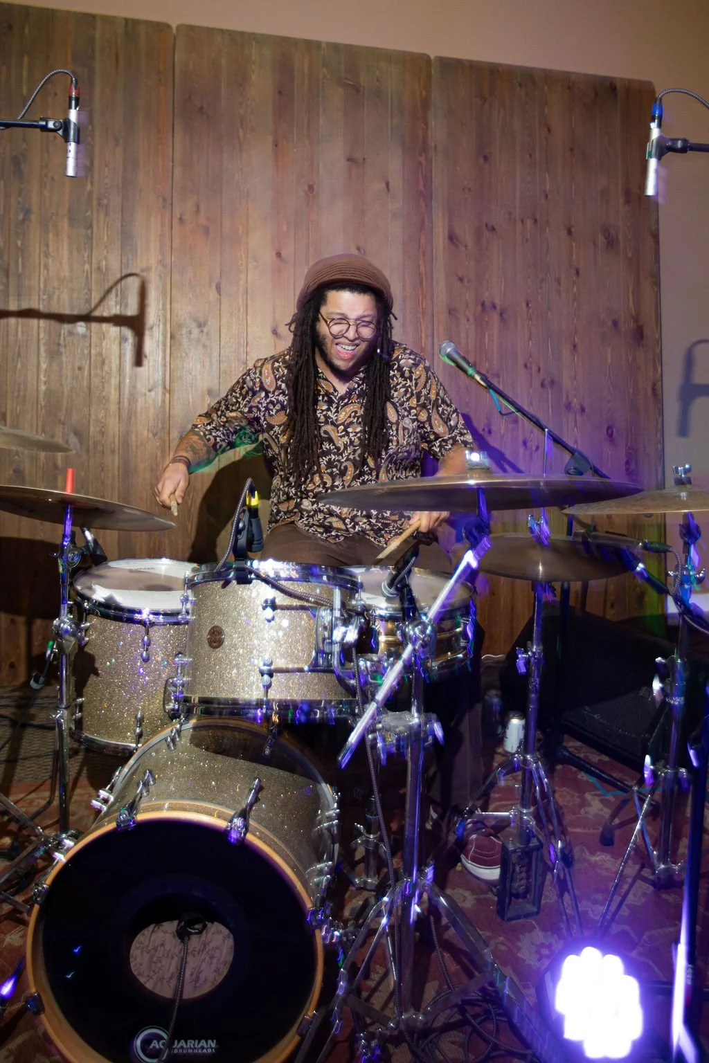 A person playing a drum set and smiling, with a wooden wall background.
