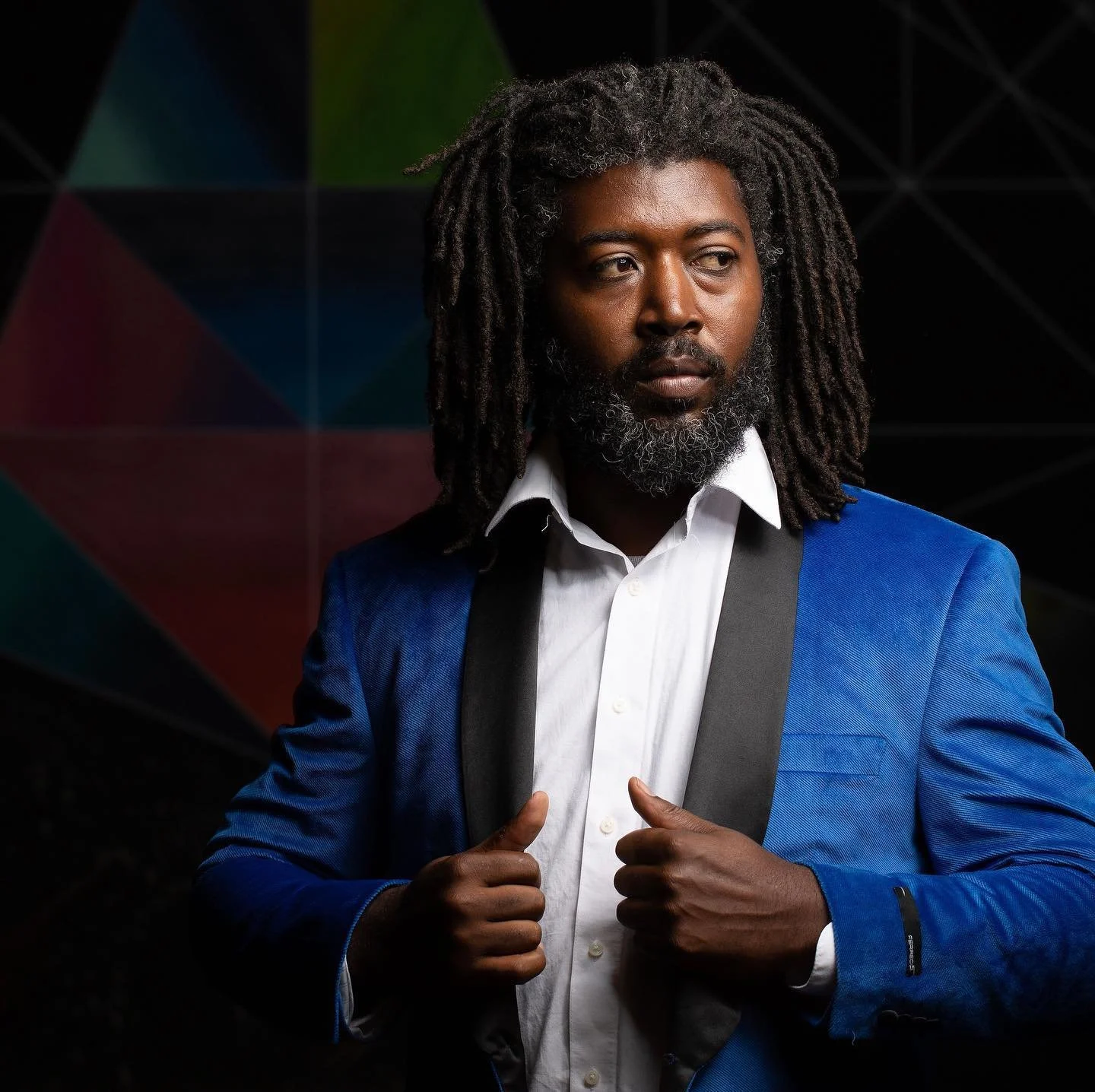 A man wearing a blue blazer and white shirt, holding the straps of a backpack, looking to the side, standing in front of a colorful, geometric background.