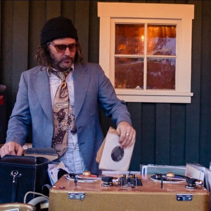 A man with long hair, a beard, glasses, a black beanie, wearing a blazer and a patterned scarf, standing behind a turntable with records, in front of a dark wood-paneled wall with a window, holding a vinyl record sleeve.