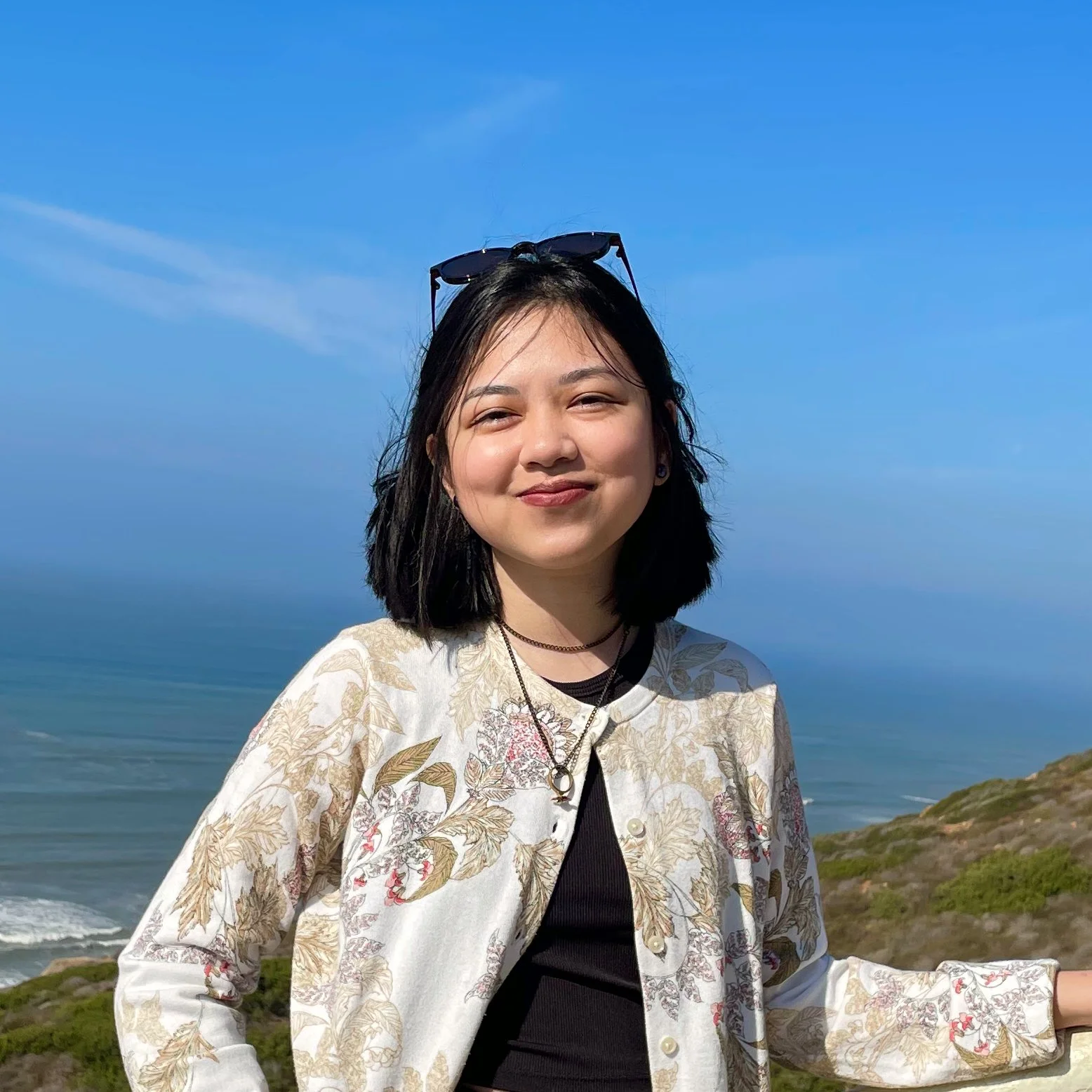 Smiling woman with black hair, wearing sunglasses on her head, a floral-patterned jacket, black top, and jewelry, standing outdoors near the coastline with blue sky and ocean in the background.