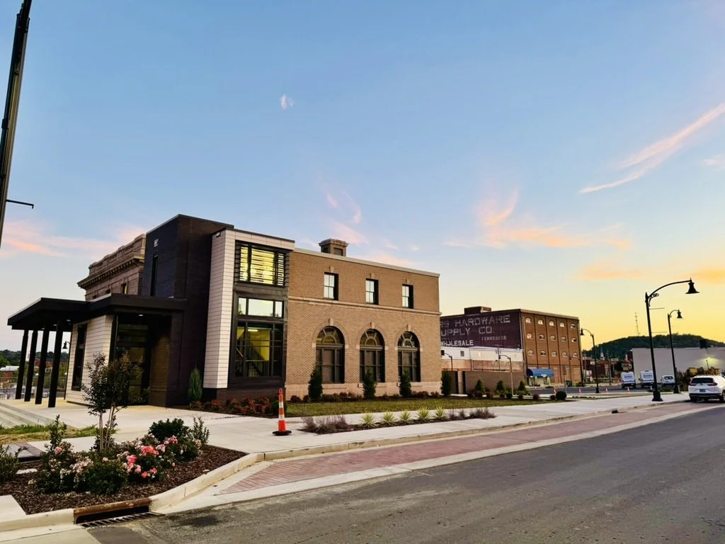 A modern commercial building with large windows and brick exterior, surrounded by a freshly landscaped sidewalk, street, and parked cars at sunset.