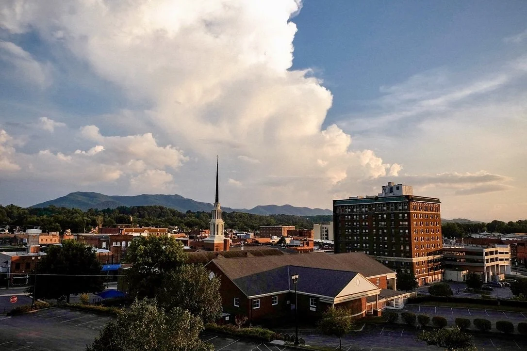 View of a city with a prominent church steeple, surrounding buildings, parking lot, and mountains in the background under a partly cloudy sky in the late afternoon or evening.