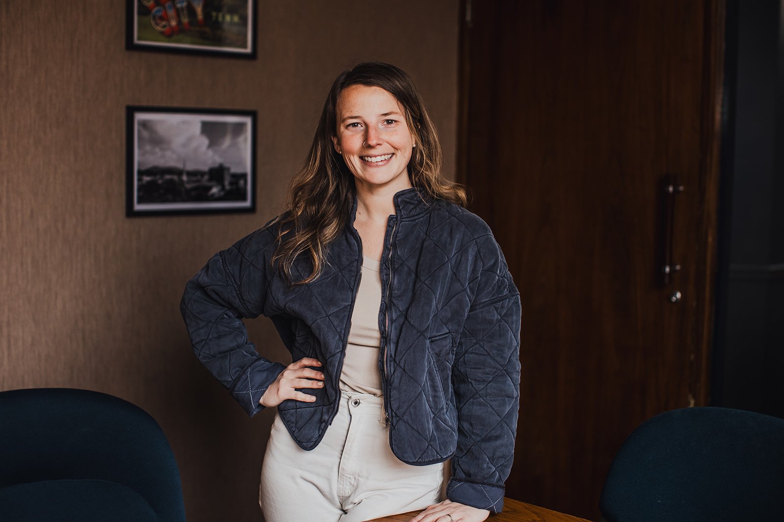 A young woman with brown hair smiling, standing in a room with brown textured walls and framed pictures hanging behind her. She is wearing a navy blue quilted jacket and beige pants, with one hand on her hip and the other resting on a table.