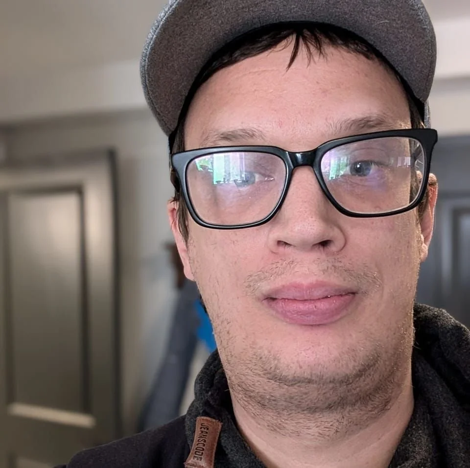 Close-up of a young man wearing glasses and a gray cap, with a slight smile, indoors.