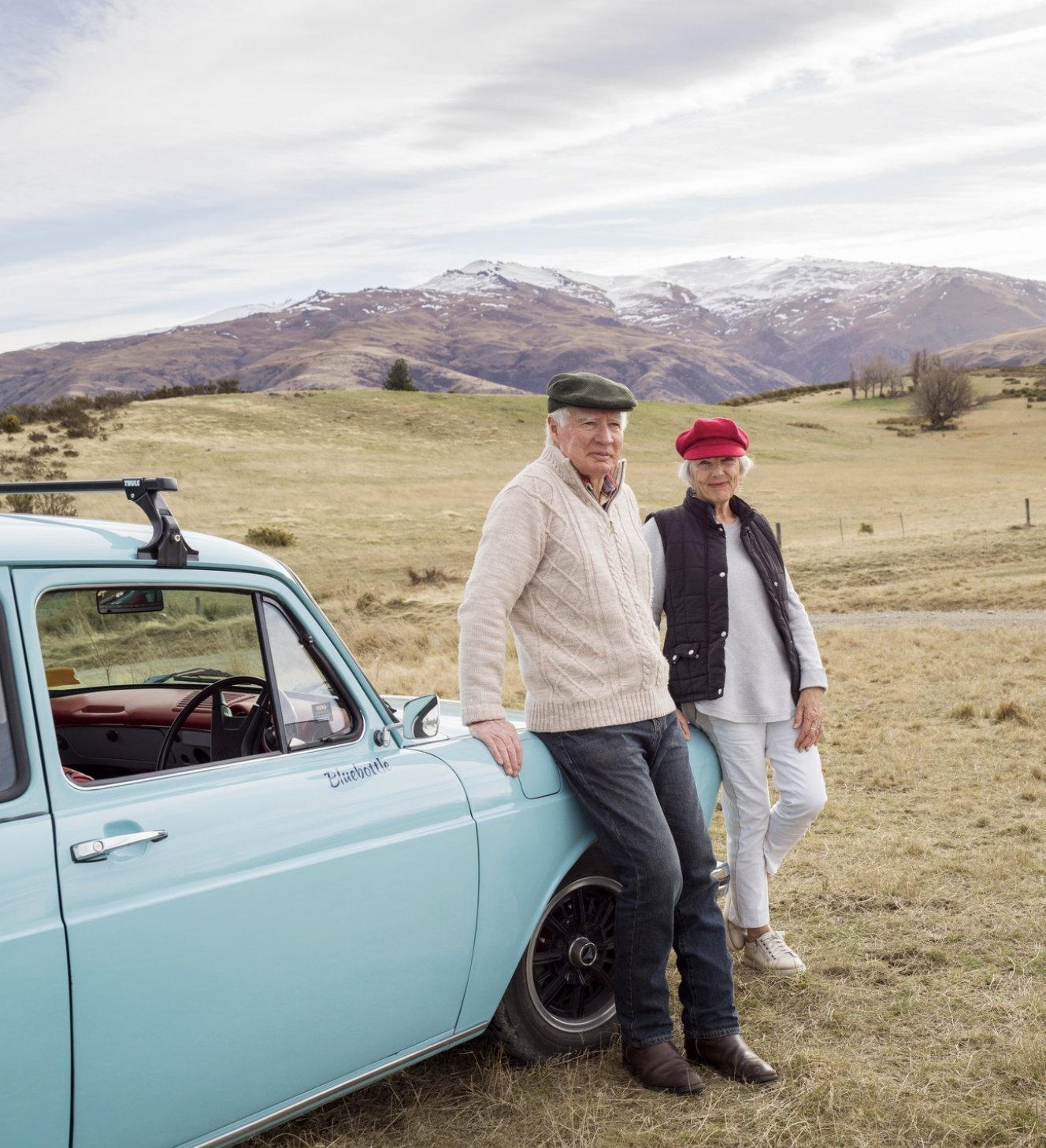 An elderly man and woman standing outdoors next to a vintage blue car with mountains in the background. The man wears a green cap and beige sweater, while the woman wears a red hat and black vest.