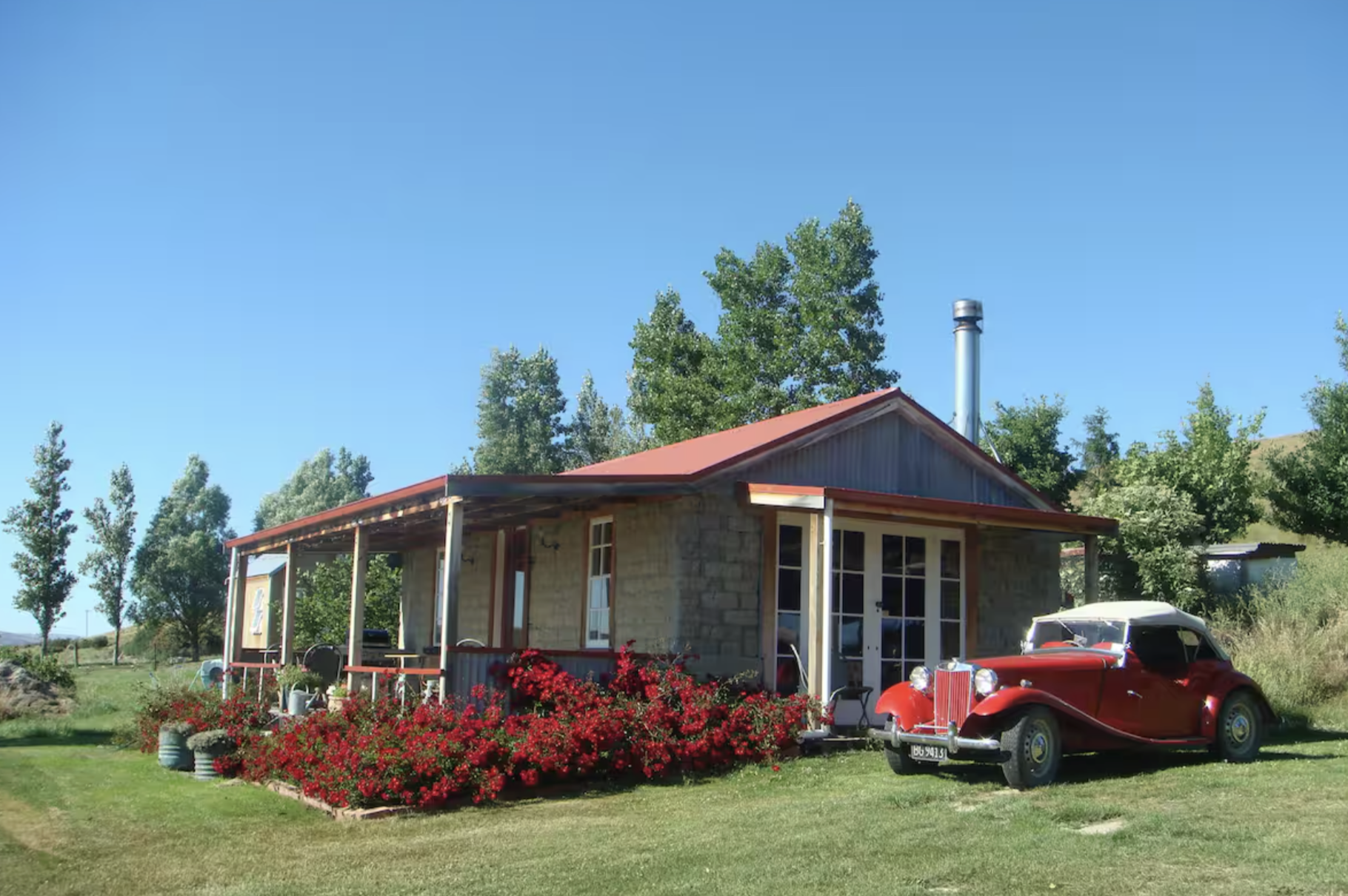 A red vintage car parked next to a rustic house with a flower garden in front, green trees, and a clear blue sky.
